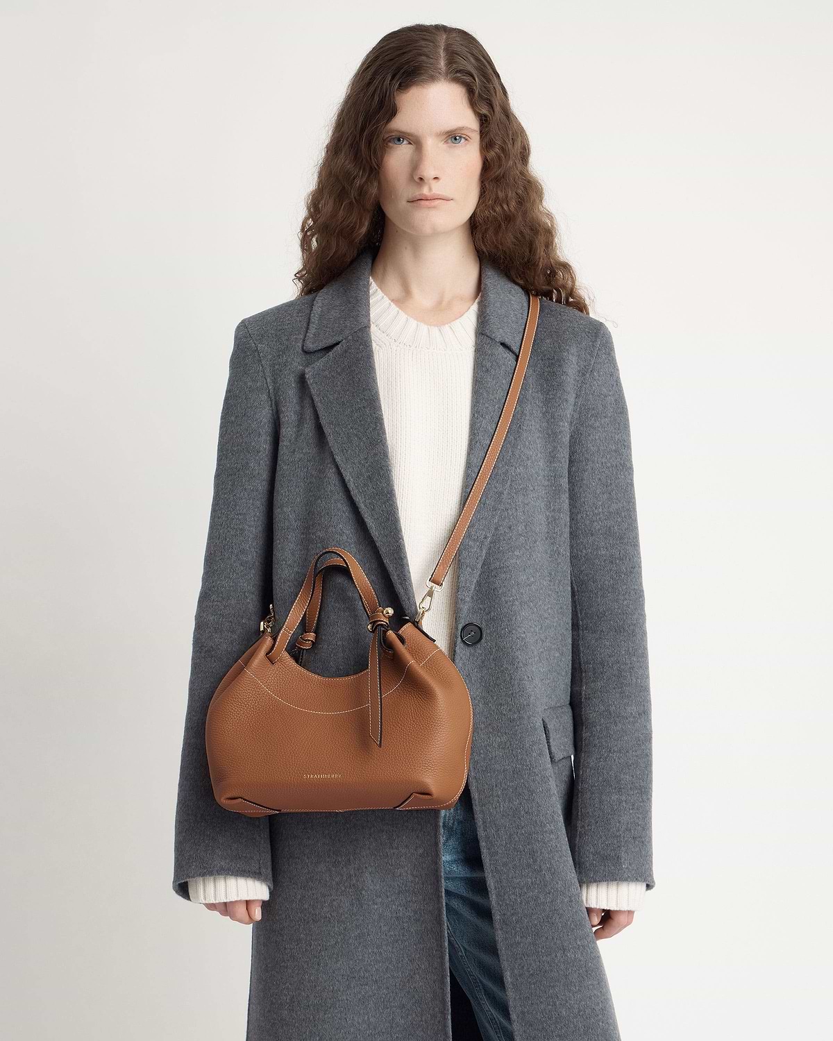 Model wearing grey coat carrying tan Strathberry Barra Mini leather crossbody bag.