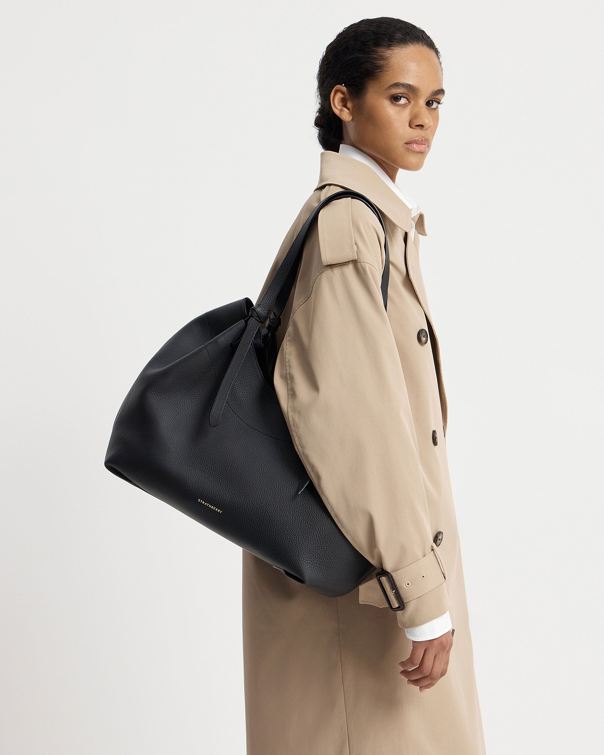 Model in a beige trench coat carrying a black Strathberry Barra tote.
