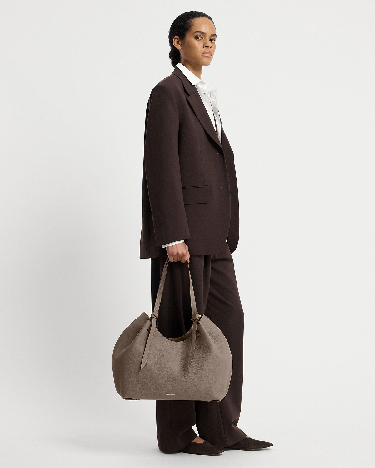 Model in dark suit holding taupe Barra Tote by Strathberry at her side.