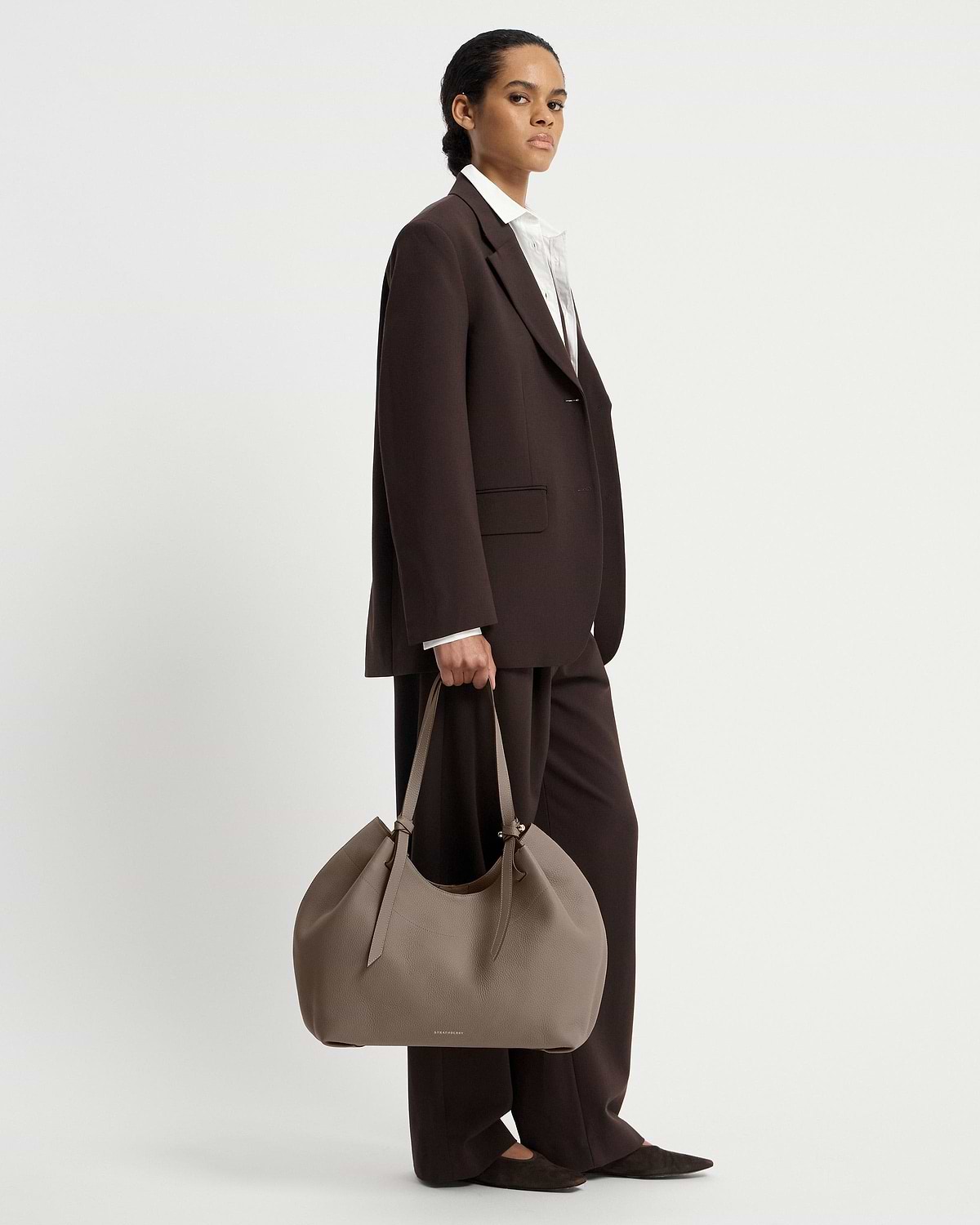 Model in dark suit holding taupe Barra Tote by Strathberry at her side.