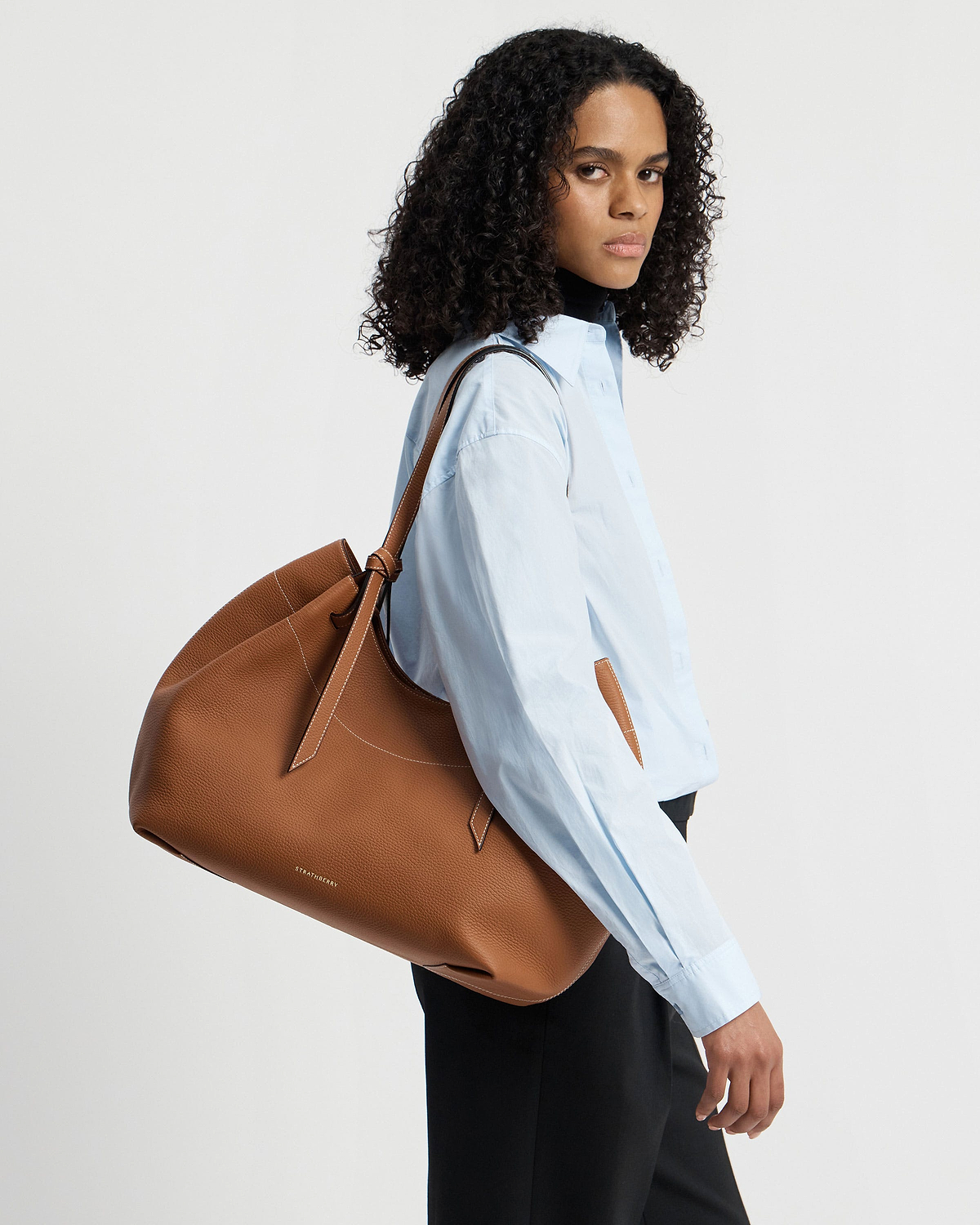 Model wearing blue shirt carries tan Strathberry Barra tote over shoulder.