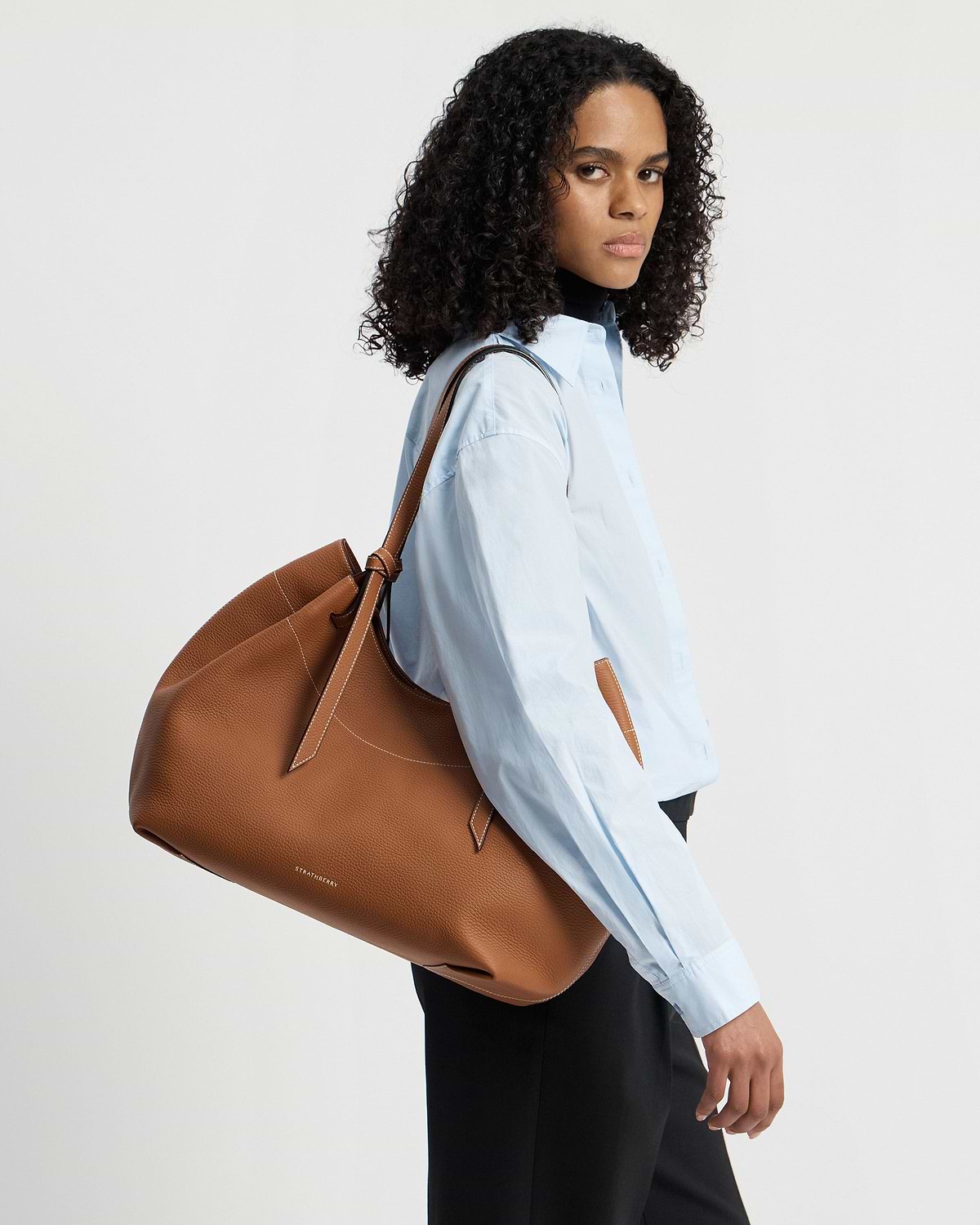 Model wearing blue shirt carries tan Strathberry Barra tote over shoulder.