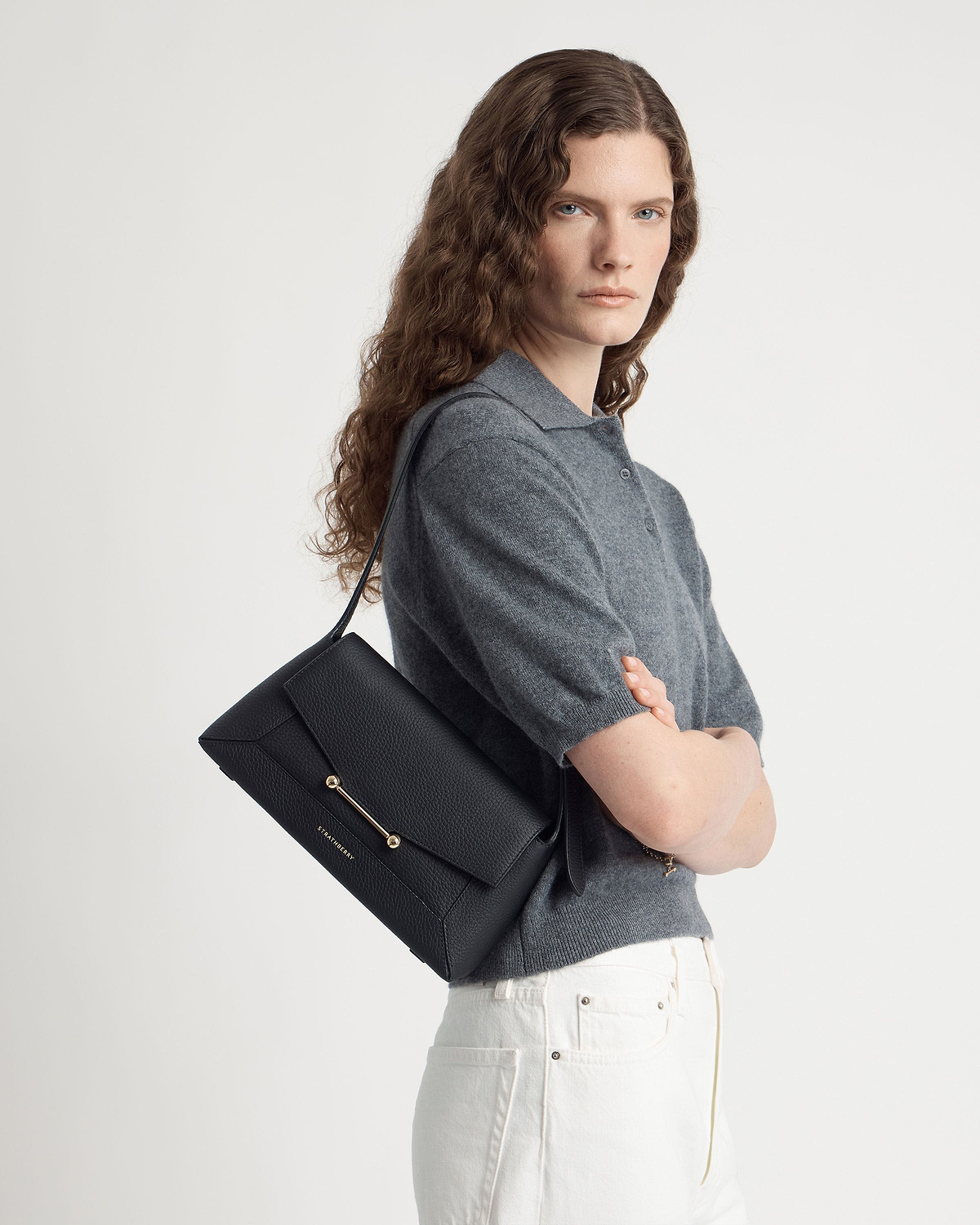 Model wearing Strathberry black mosaic shoulder bag over shoulder, grey top, white jeans.