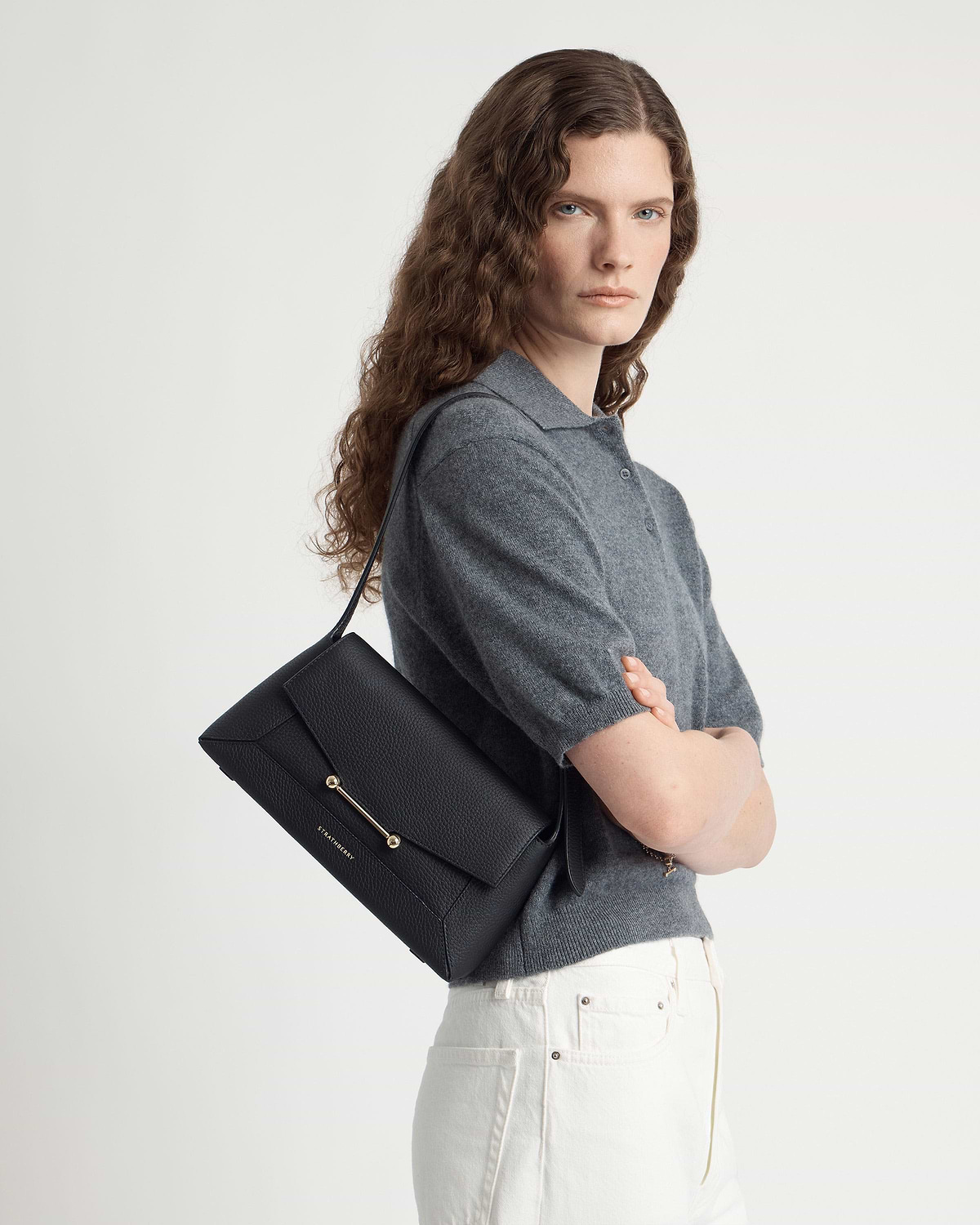 Model wearing Strathberry black mosaic shoulder bag over shoulder, grey top, white jeans.