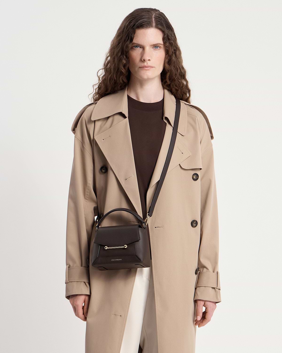 Model wearing trench coat with small Strathberry Mosaic Nano espresso leather top-handle bag.