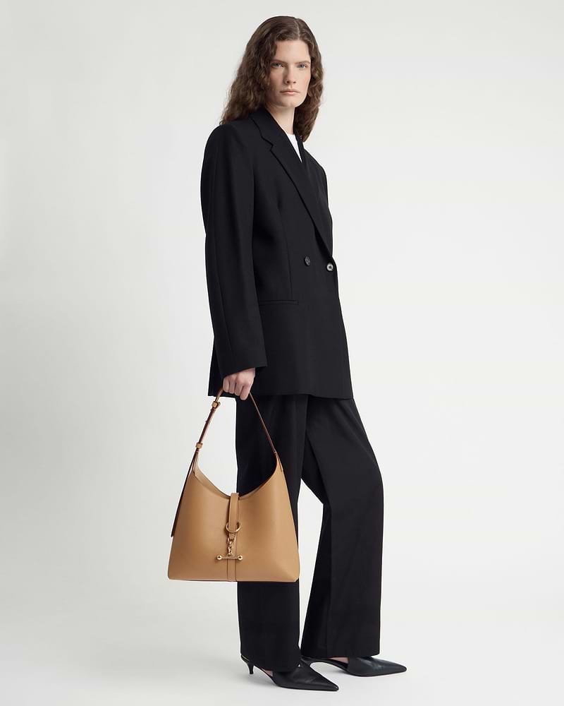 Model in black suit holding caramel Kite Hobo bag by top strap.