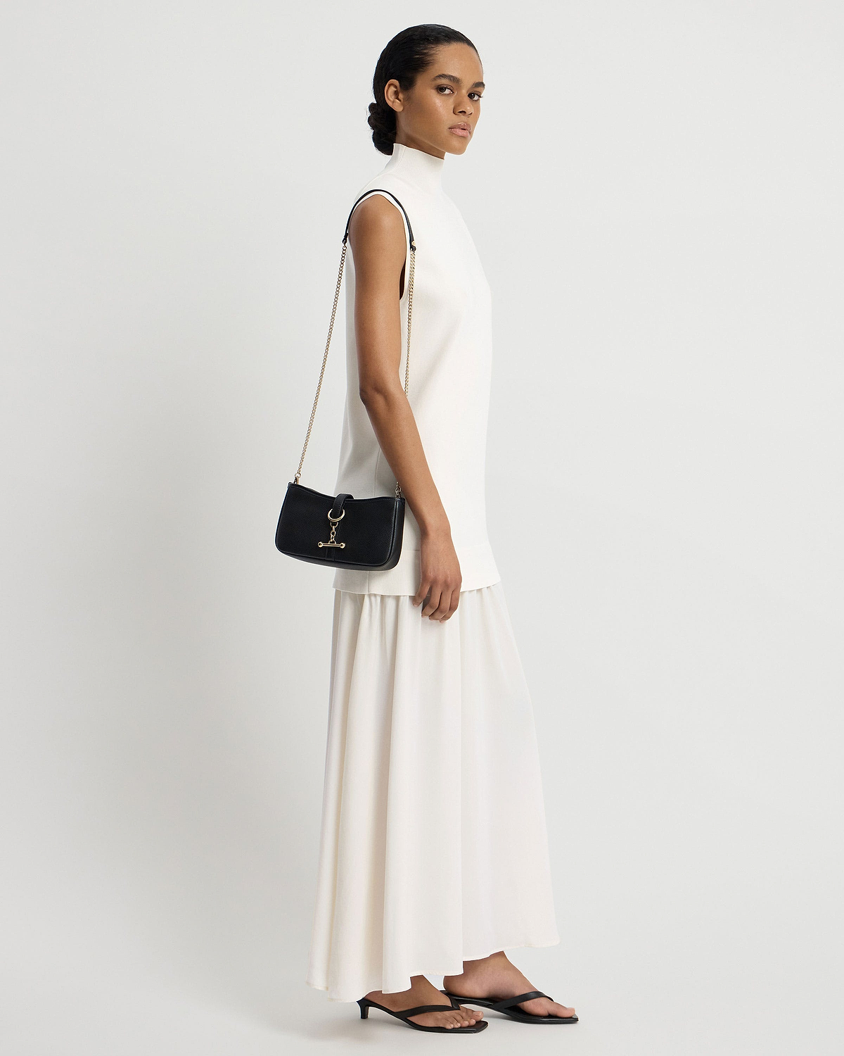 Model wearing white sleeveless dress holds black shoulder bag with gold chain strap.