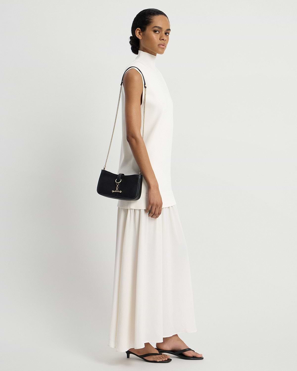 Model wearing white sleeveless dress holds black shoulder bag with gold chain strap.
