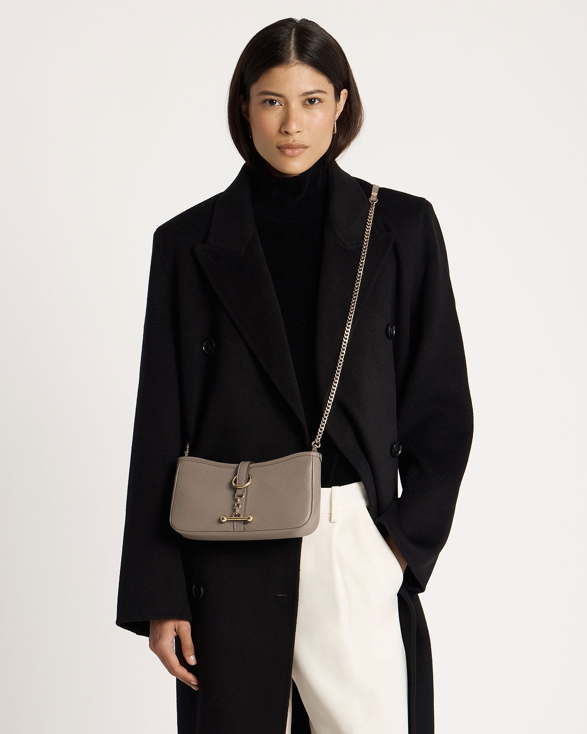 Model wearing black coat and turtleneck with taupe crossbody bag on gold chain.