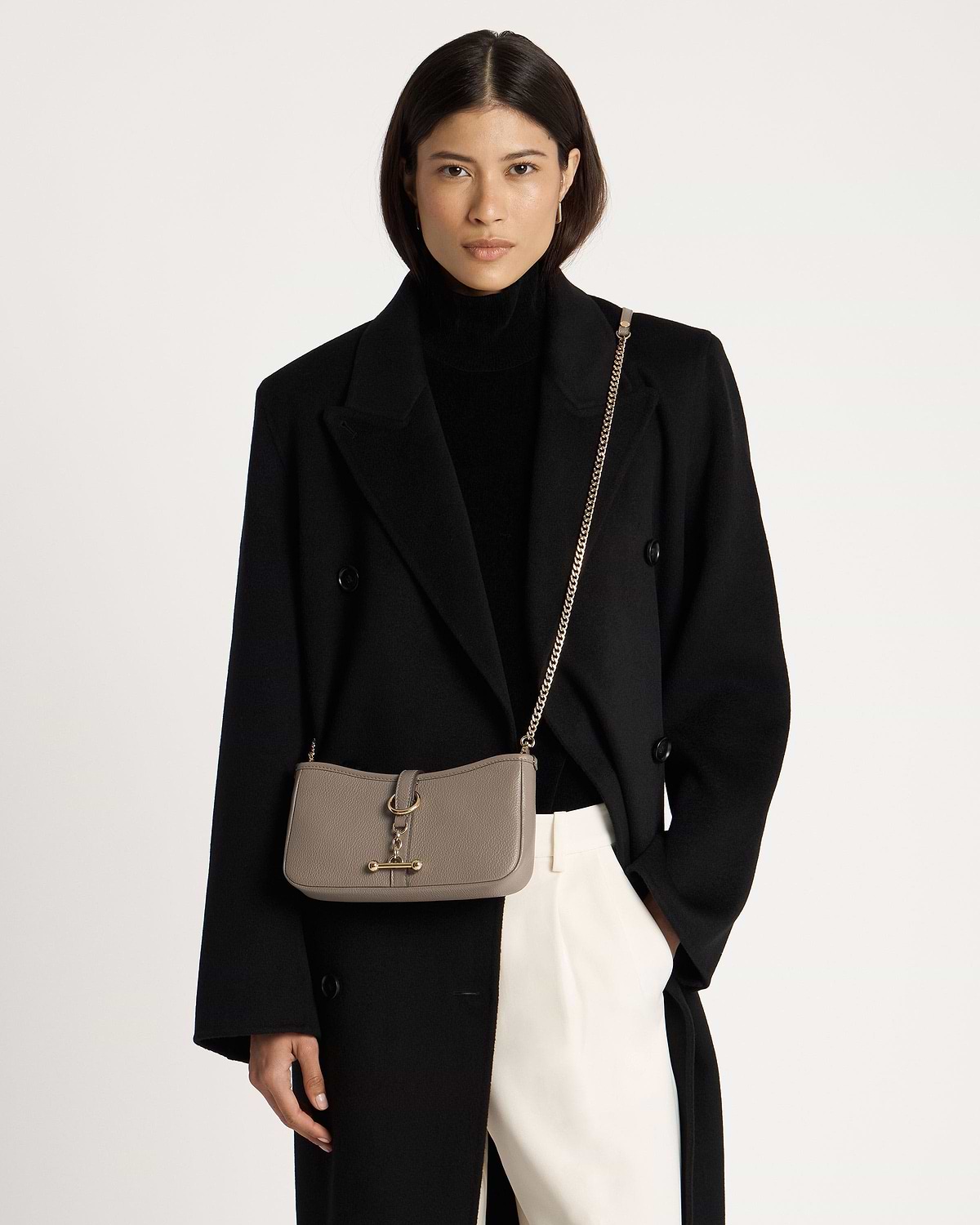 Model wearing black coat and turtleneck with taupe crossbody bag on gold chain.