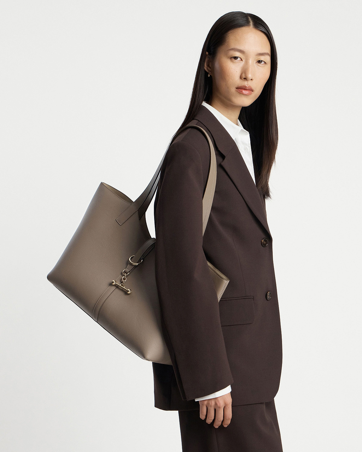 Woman wearing a dark brown suit carrying a taupe leather Kite Tote on her shoulder.
