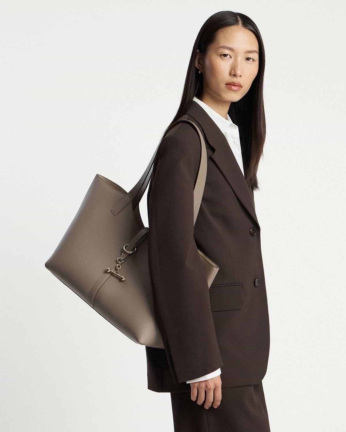 Woman wearing a dark brown suit carrying a taupe leather Kite Tote on her shoulder.