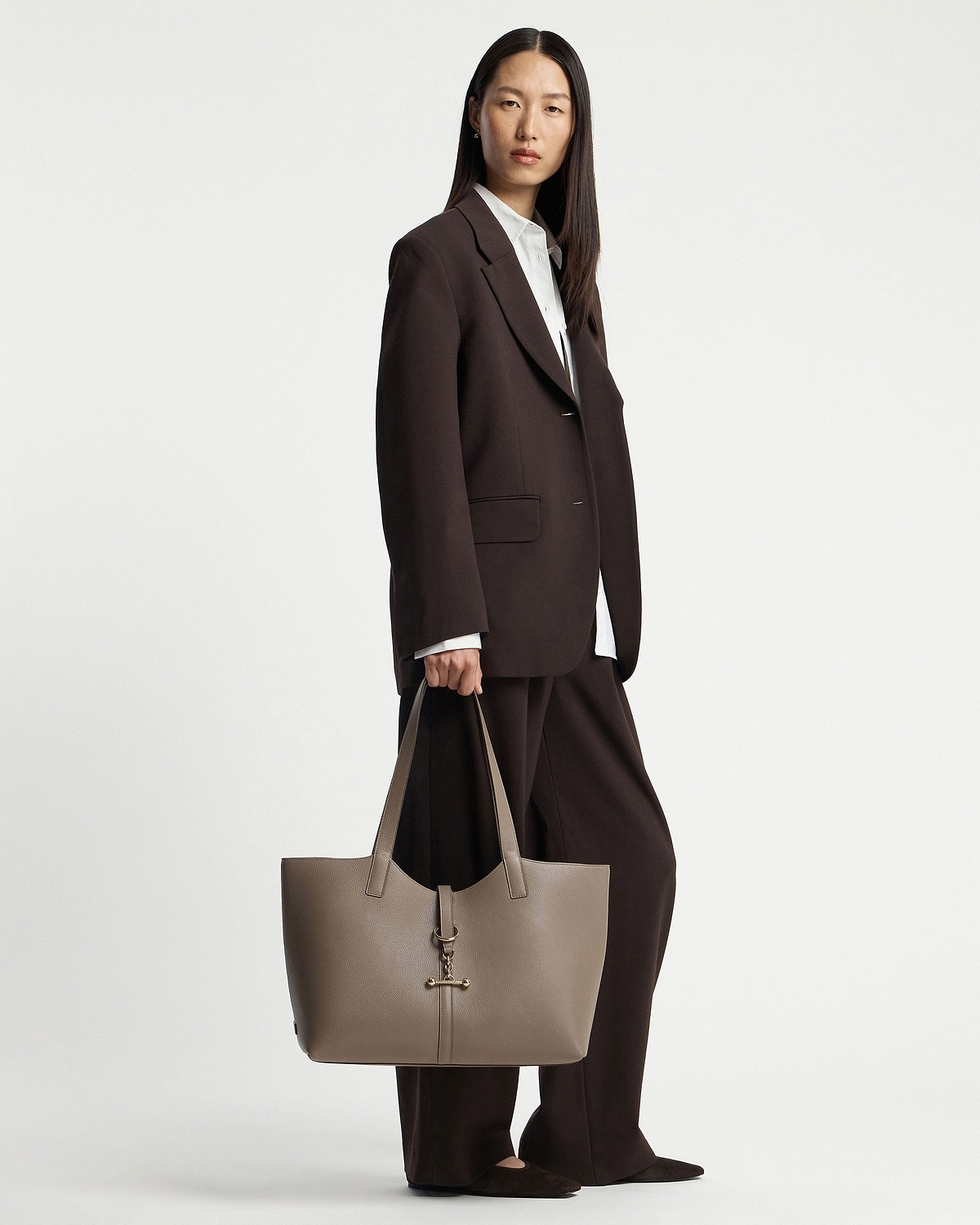 Model standing in dark brown suit holding a taupe tote bag with gold hardware.