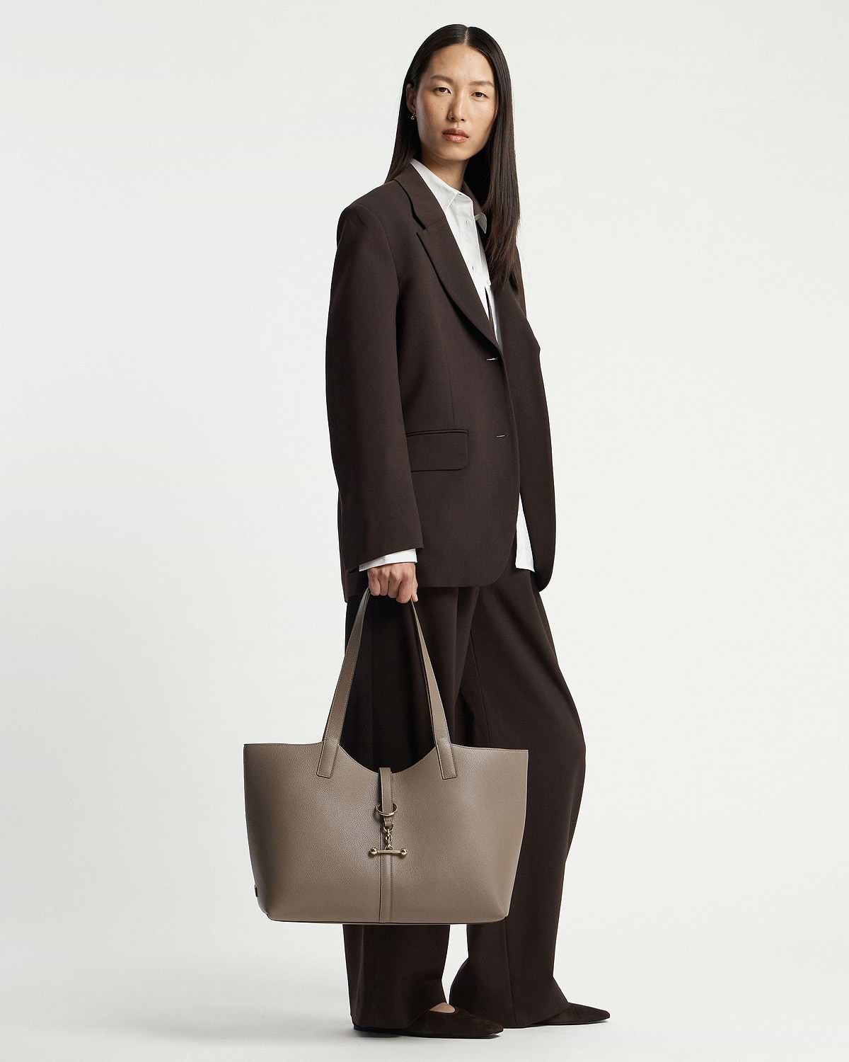 Model standing in dark brown suit holding a taupe tote bag with gold hardware.