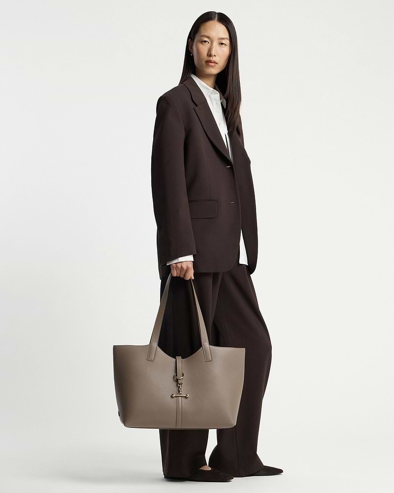 Model standing in dark brown suit holding a taupe tote bag with gold hardware.