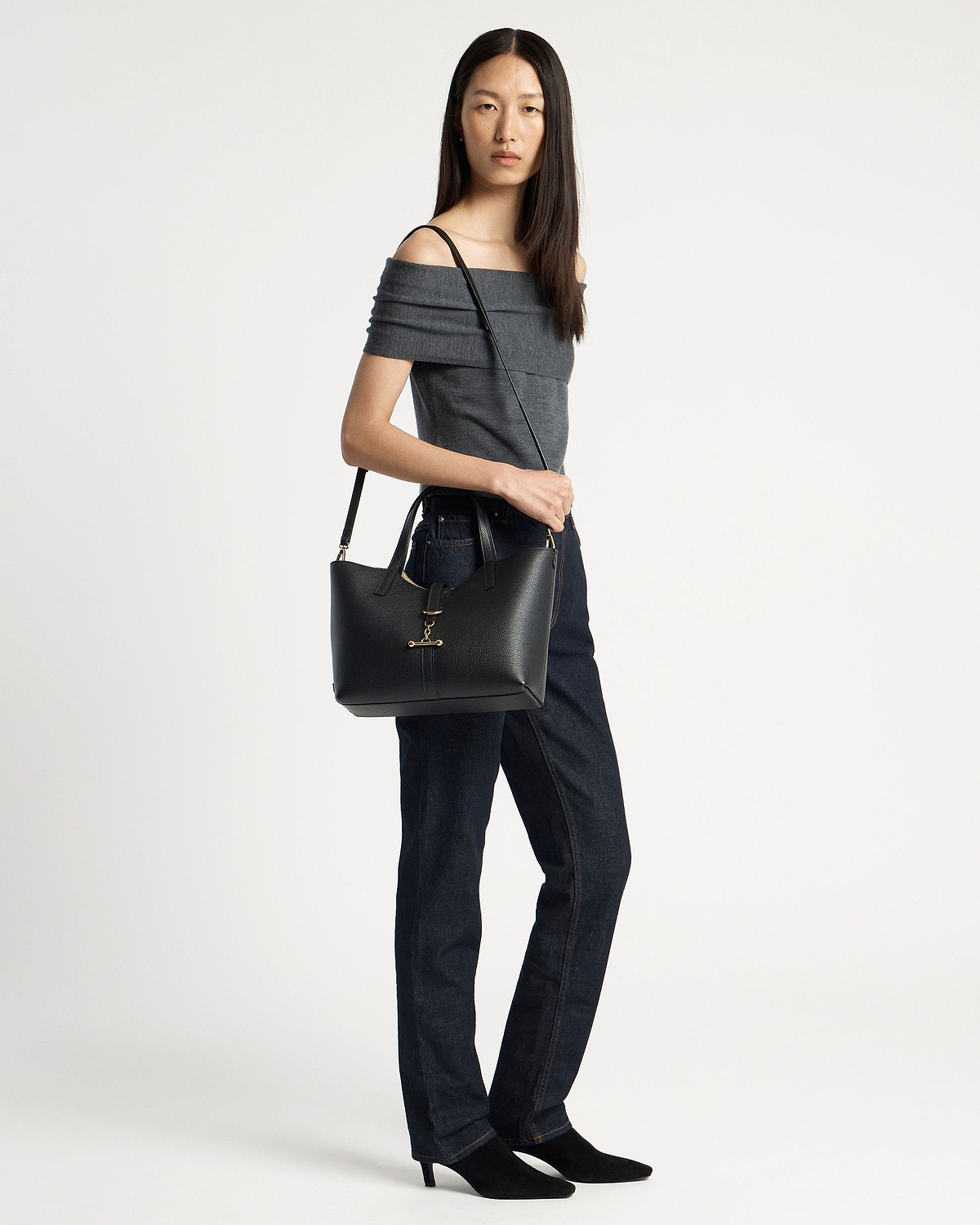 Model wearing grey off-shoulder top and dark jeans carrying black tote bag.