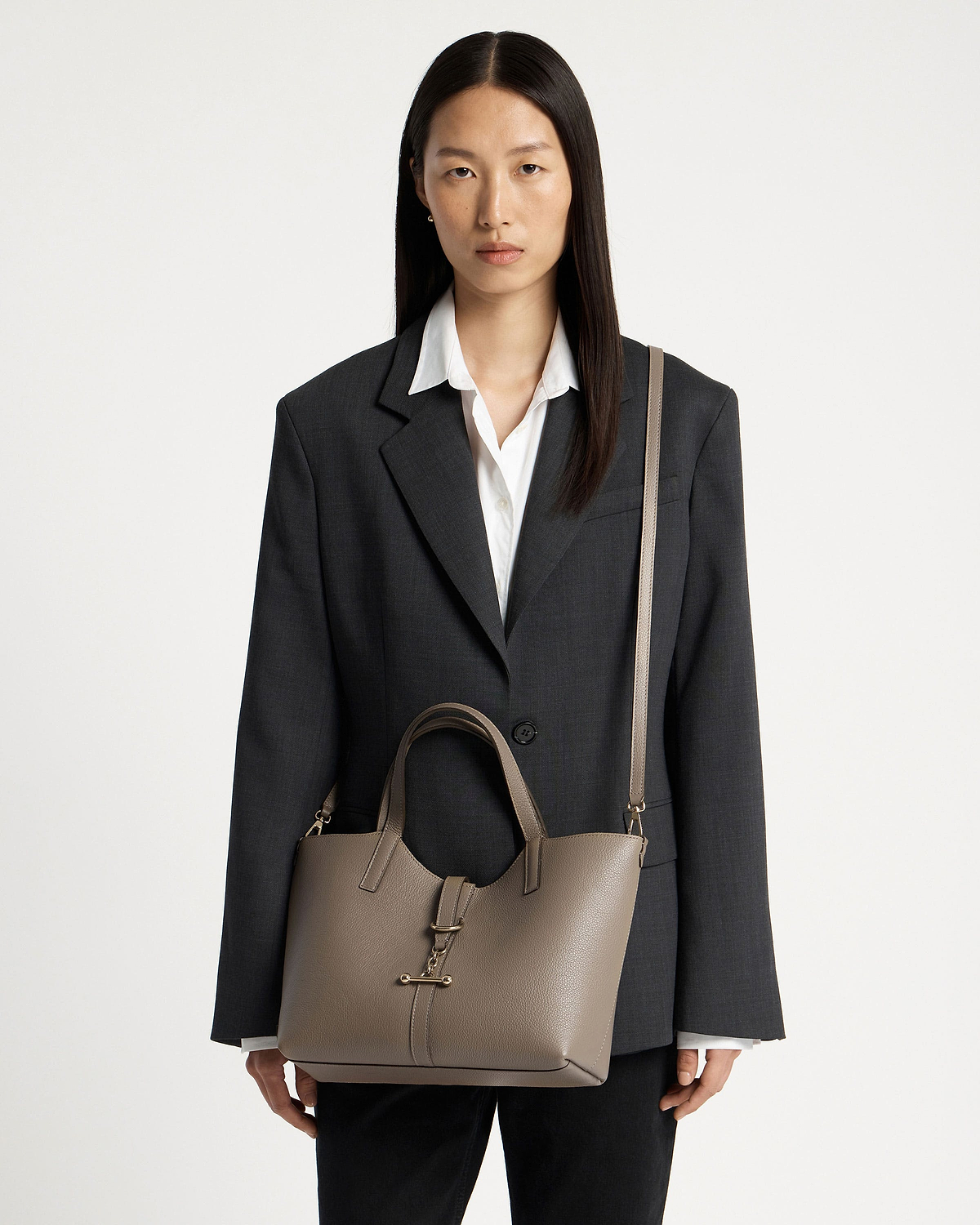 Model wearing a dark blazer carrying a taupe leather tote bag with shoulder strap