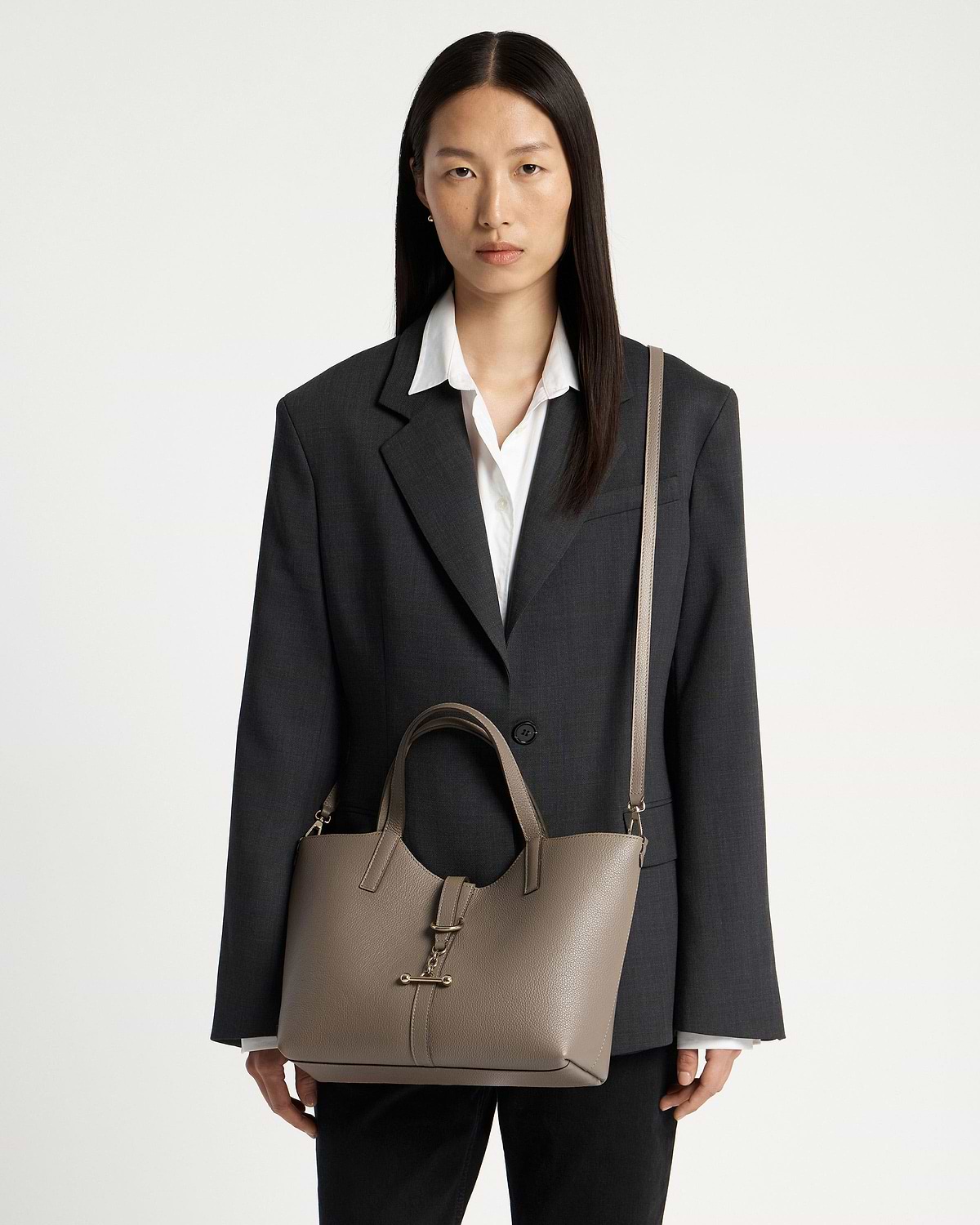 Model wearing a dark blazer carrying a taupe leather tote bag with shoulder strap