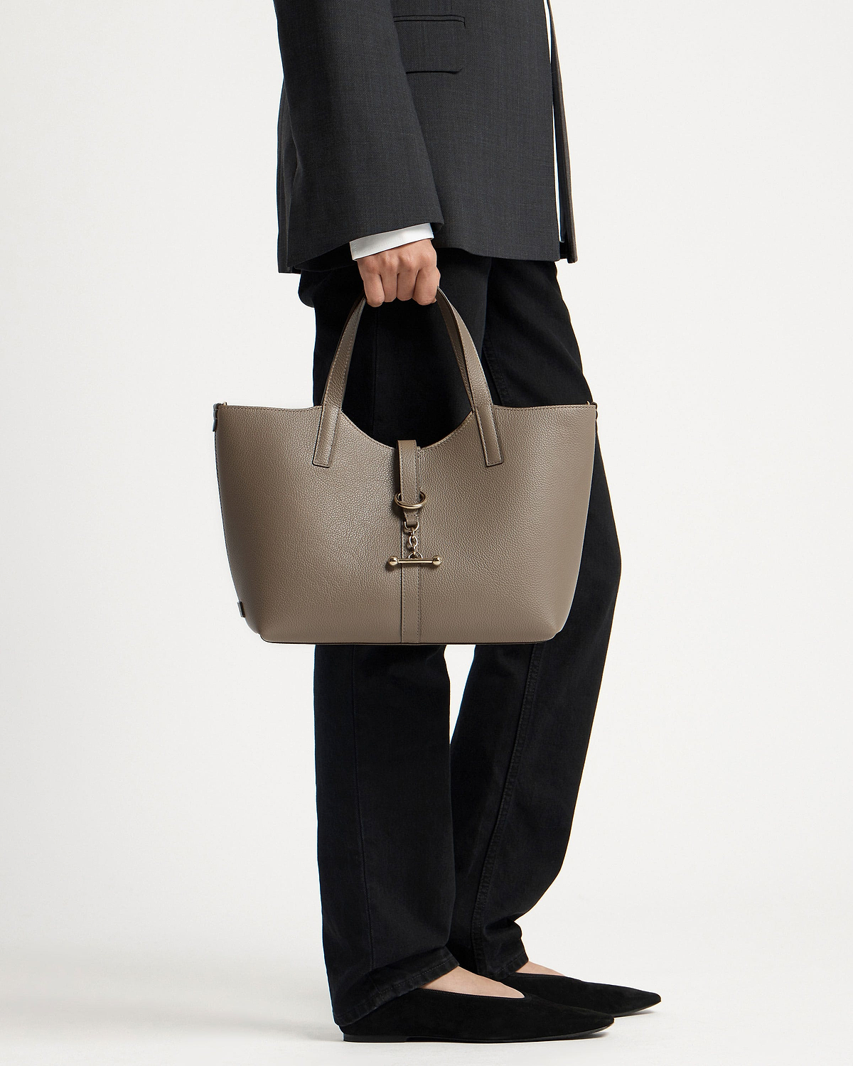 Model holding taupe Kite Tote Midi with metal bar charm, wearing black trousers.