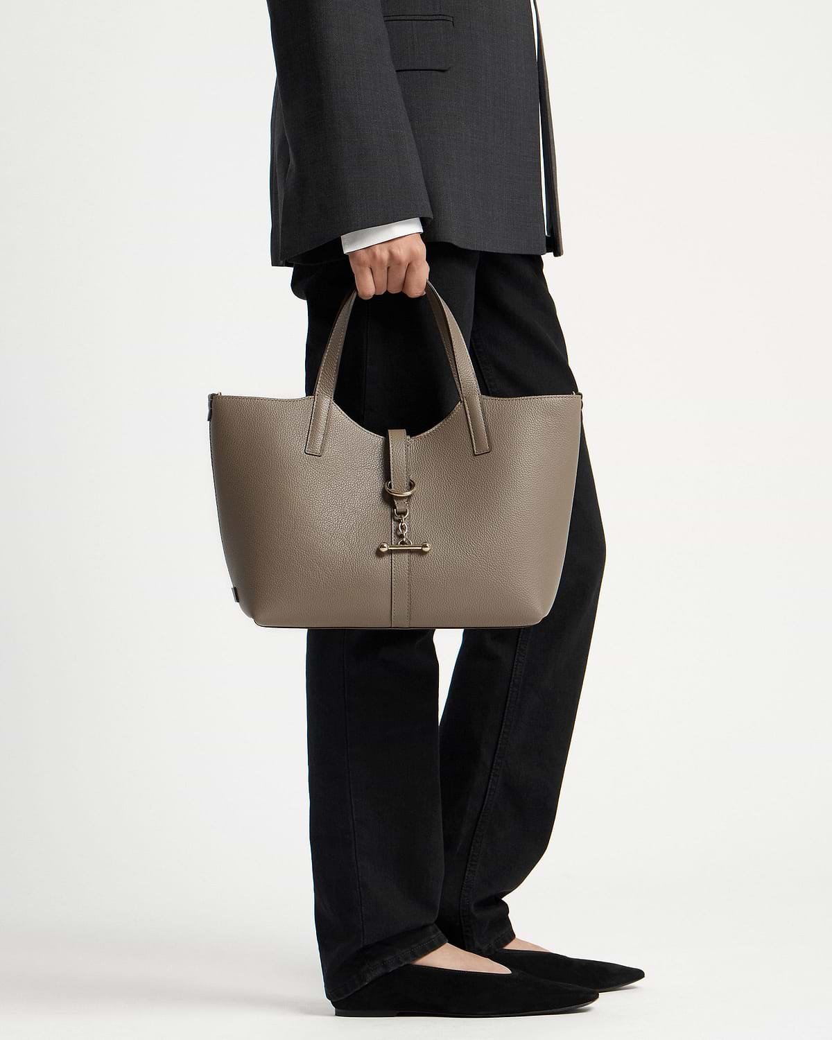 Model holding taupe Kite Tote Midi with metal bar charm, wearing black trousers.