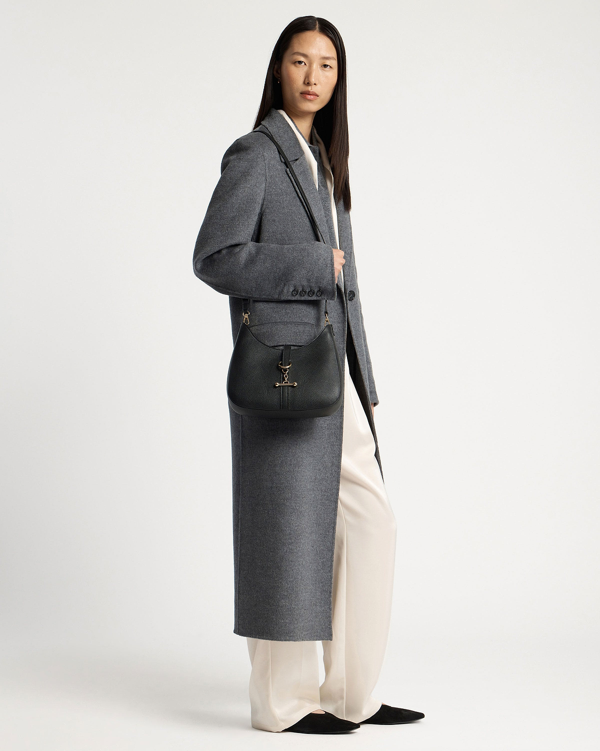 Model wearing long grey coat carrying a black kite sling bag with gold hardware.