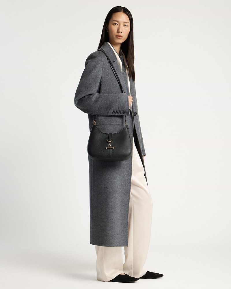 Model wearing long grey coat carrying a black kite sling bag with gold hardware.