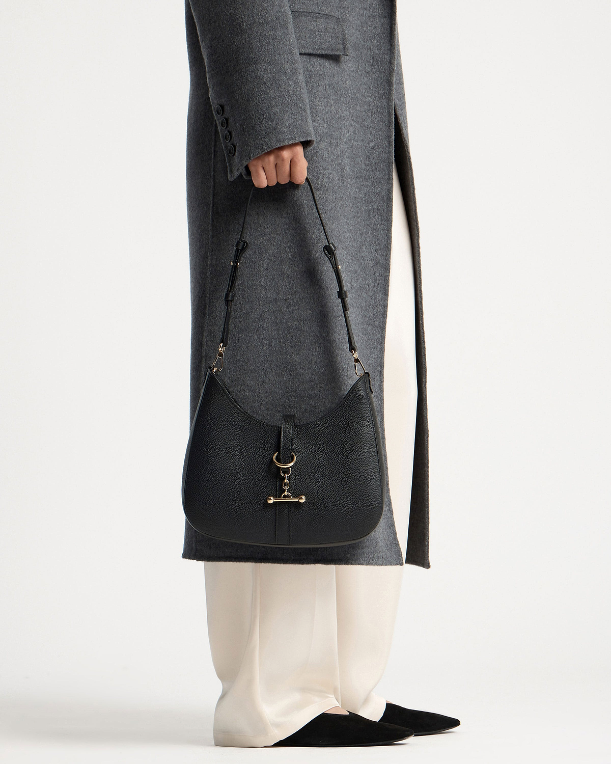 Black Kite Sling with gold hardware held beside grey coat and white trousers.