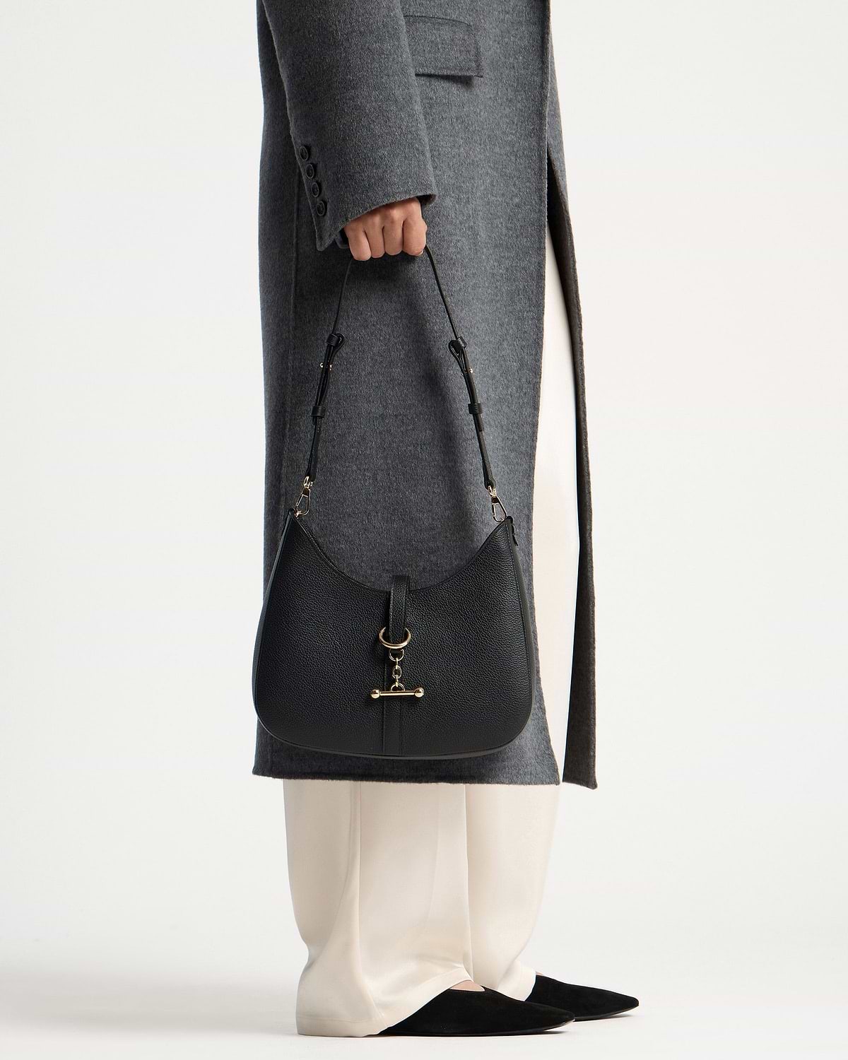 Black Kite Sling with gold hardware held beside grey coat and white trousers.
