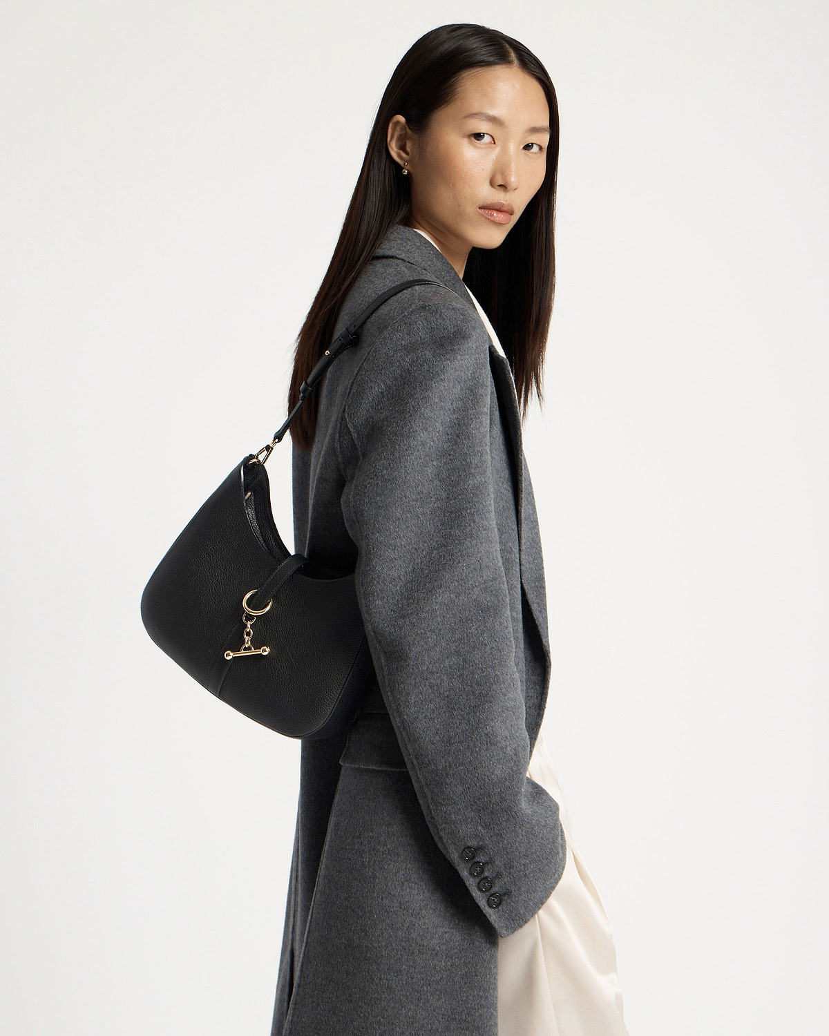 Woman wearing black kite sling bag with gold hardware over shoulder in grey coat