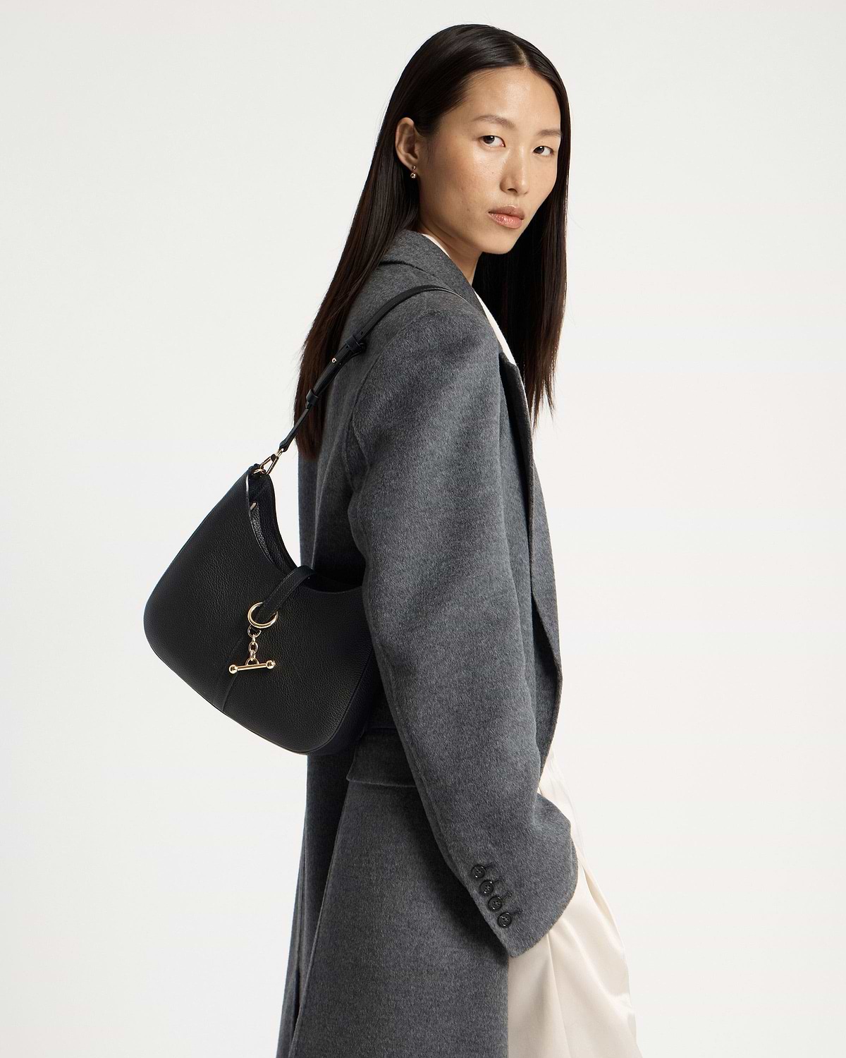 Woman wearing black kite sling bag with gold hardware over shoulder in grey coat