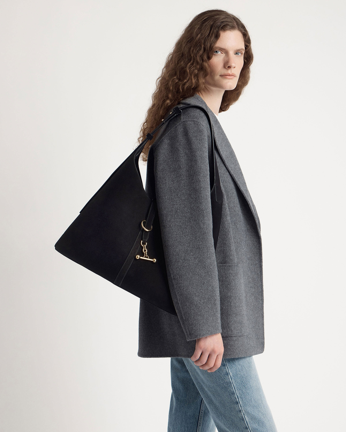 Model in grey coat and jeans carrying a large black suede hobo shoulder bag.