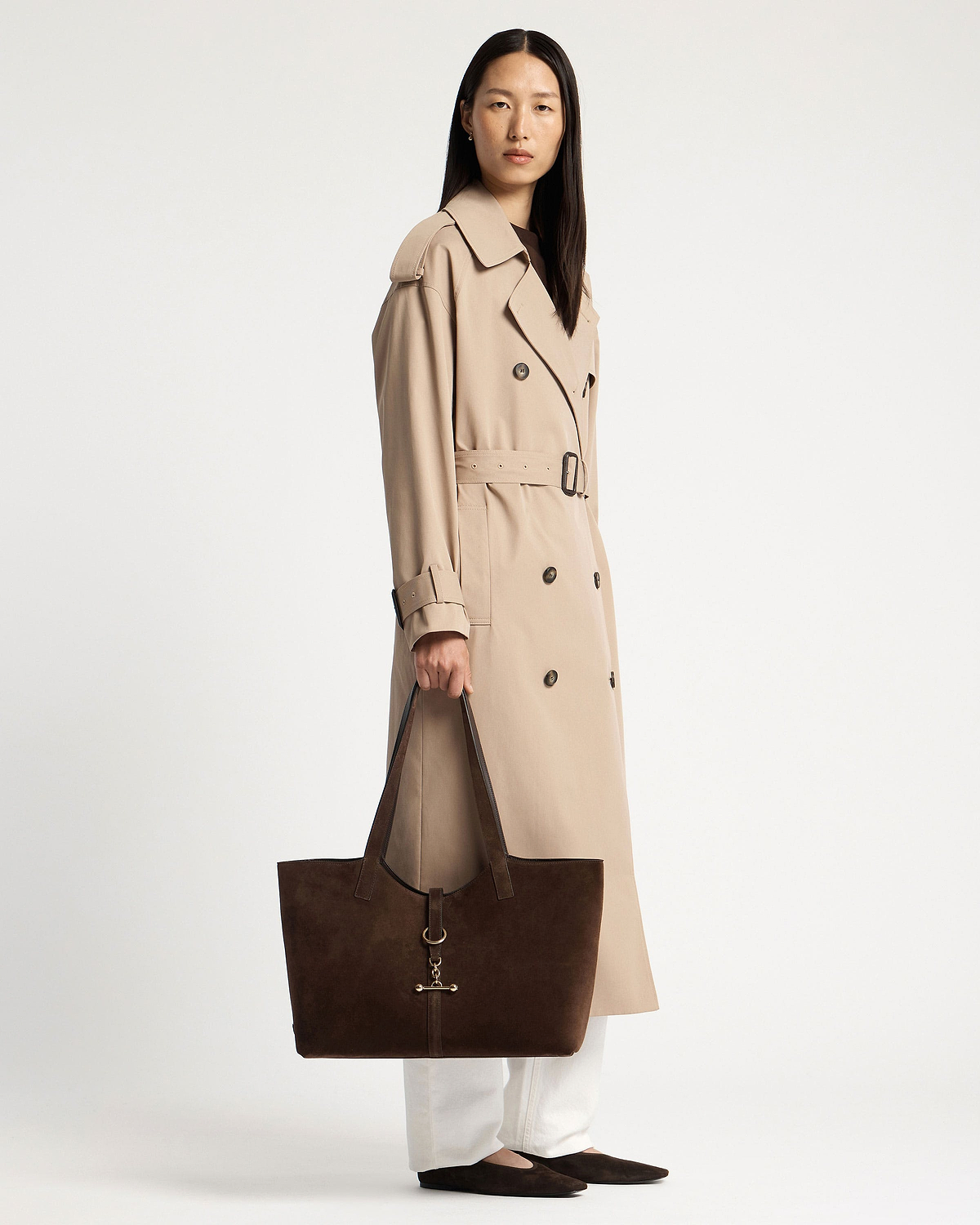 Woman in beige trench holding chocolate suede Kite tote with gold hardware.