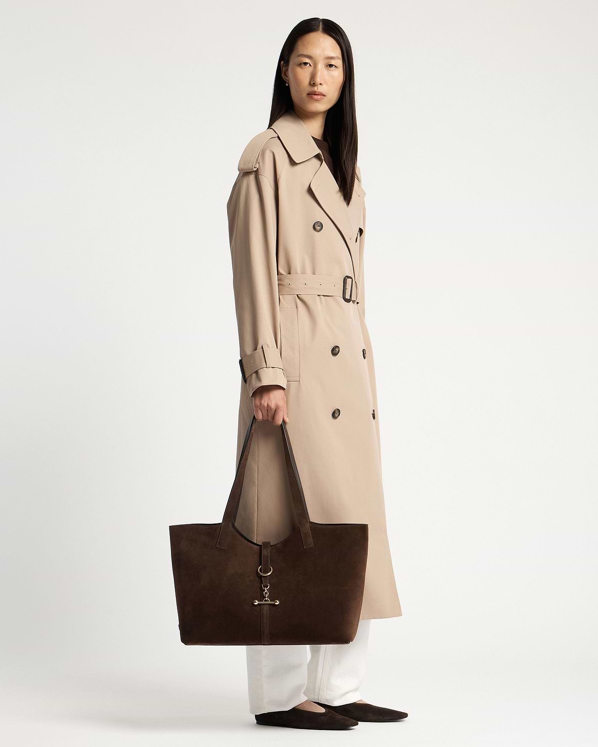 Woman in beige trench holding chocolate suede Kite tote with gold hardware.