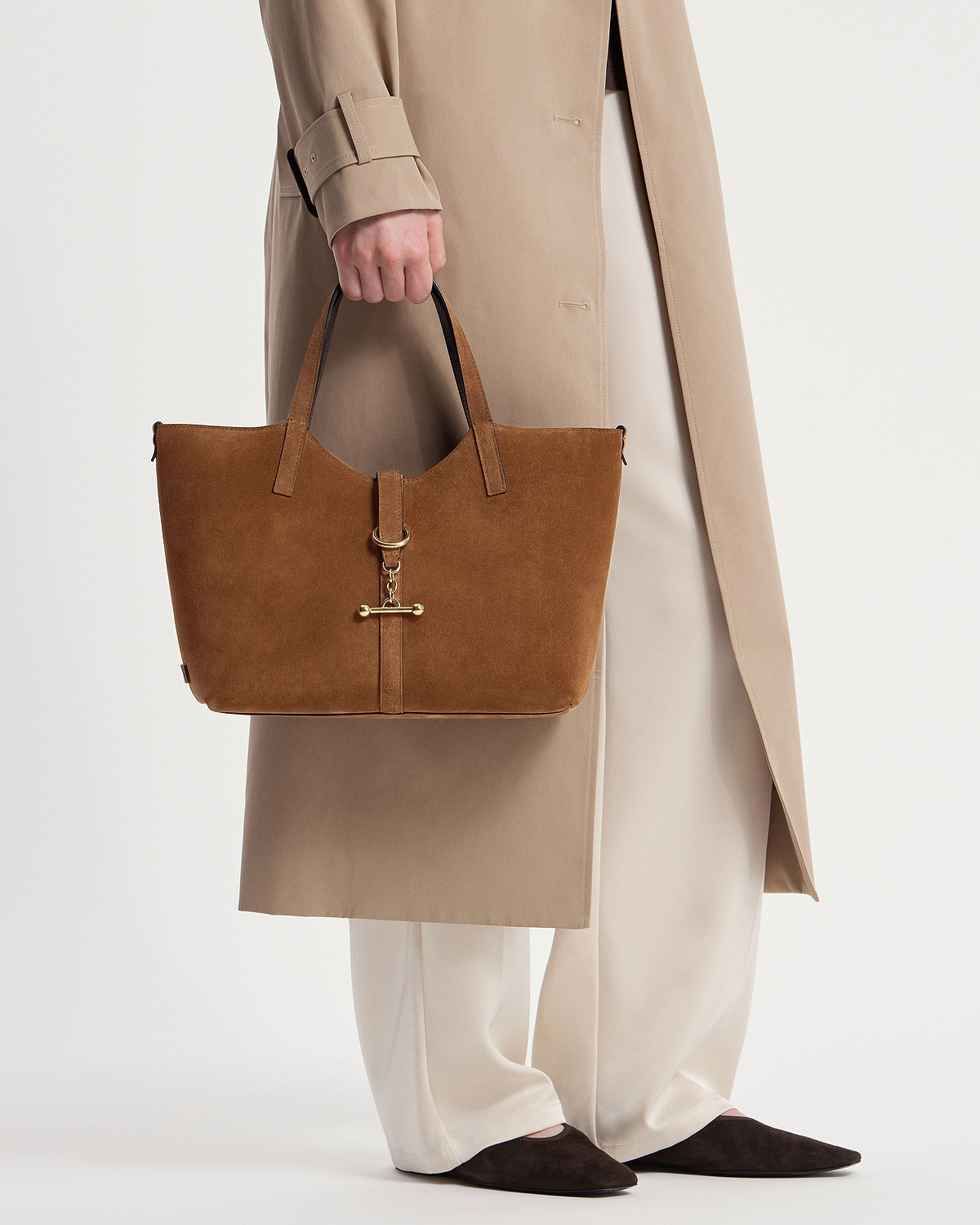 Tan suede tote bag with gold hardware held by a model wearing a beige coat.