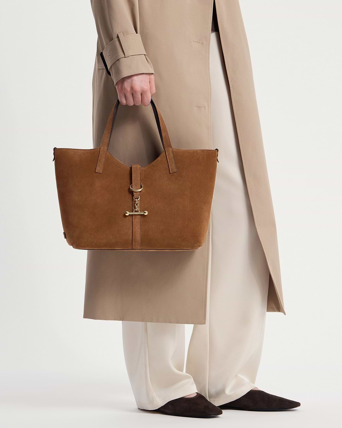 Tan suede tote bag with gold hardware held by a model wearing a beige coat.