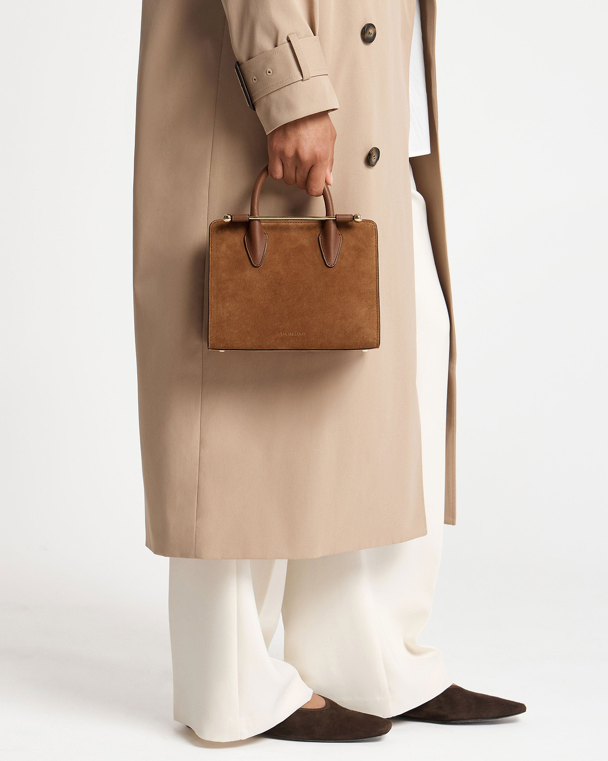 Hand holding tan suede mini tote with beige coat and white trousers.