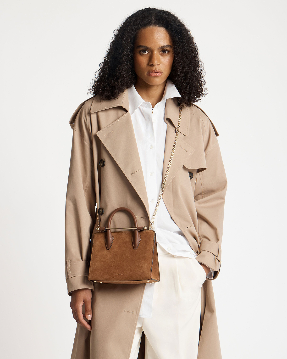 Model wearing trench coat carrying tan suede mini tote with top handles and chain strap.
