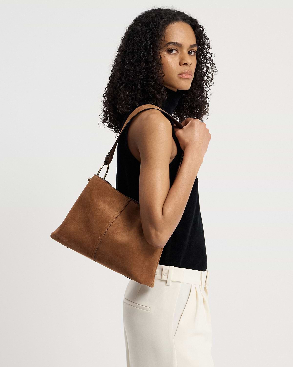 Woman wearing black sleeveless top carrying tan suede hobo bag on her shoulder.