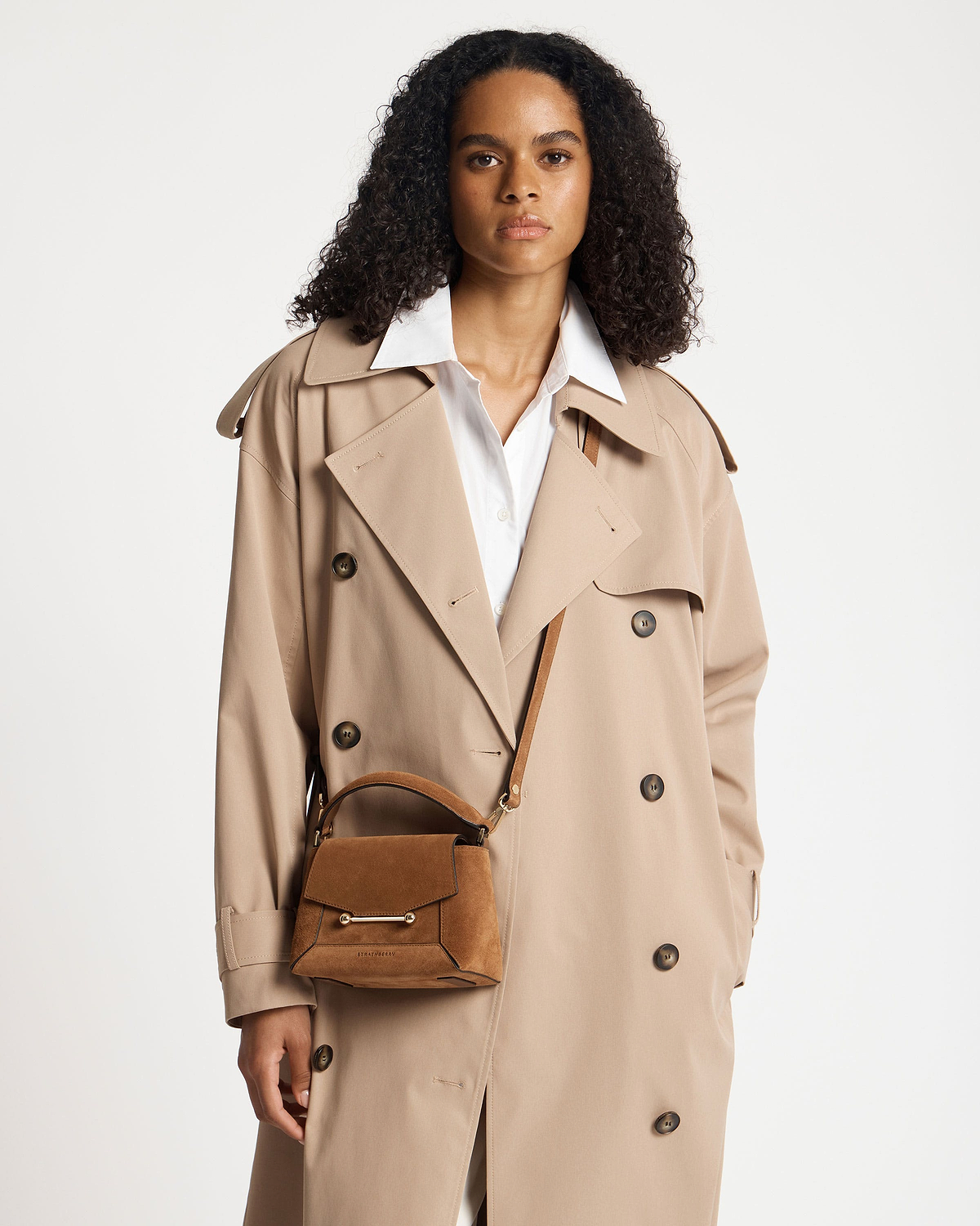 Woman wearing tan suede Mosaic Nano bag by Strathberry and a beige trench coat.