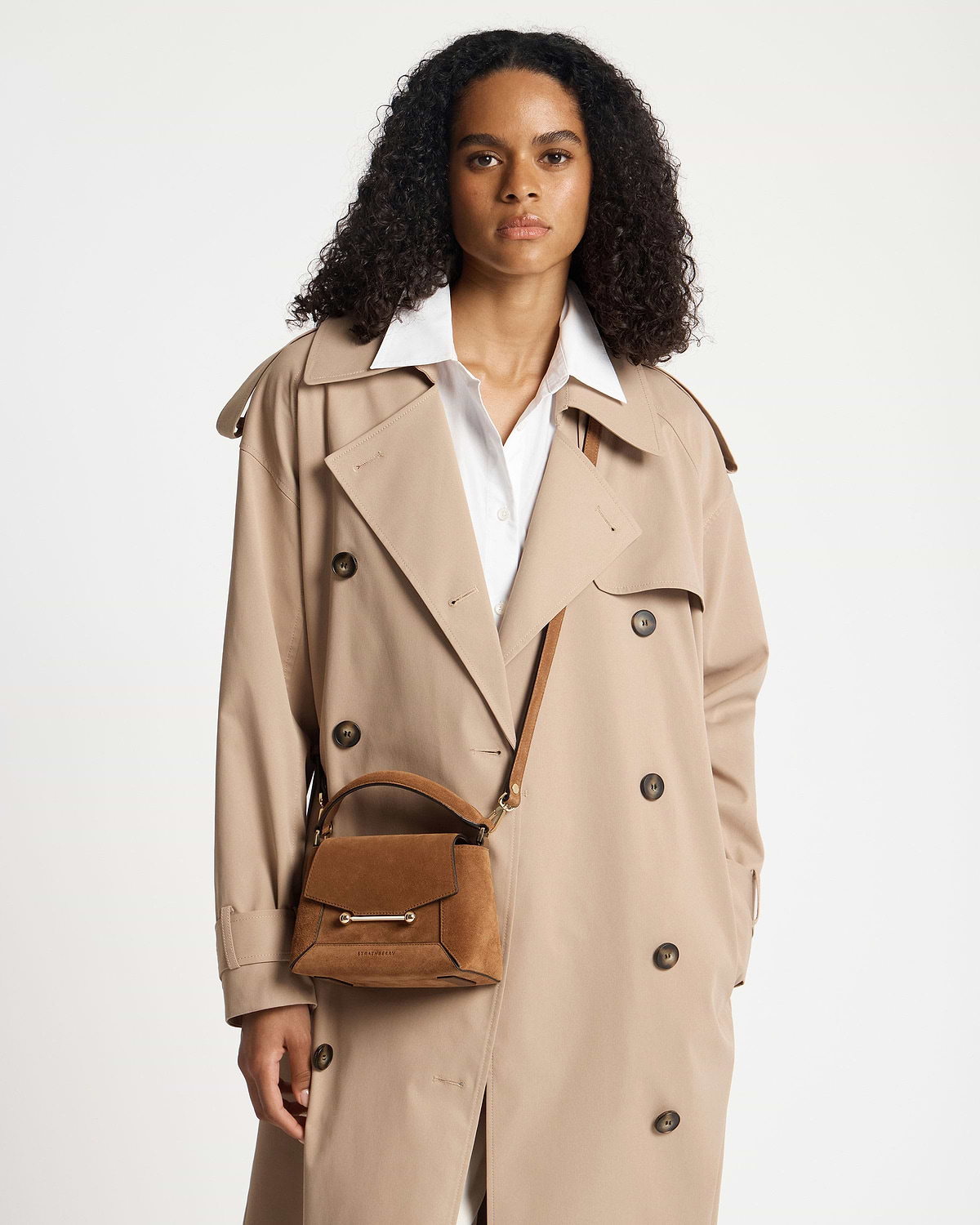 Woman wearing tan suede Mosaic Nano bag by Strathberry and a beige trench coat.