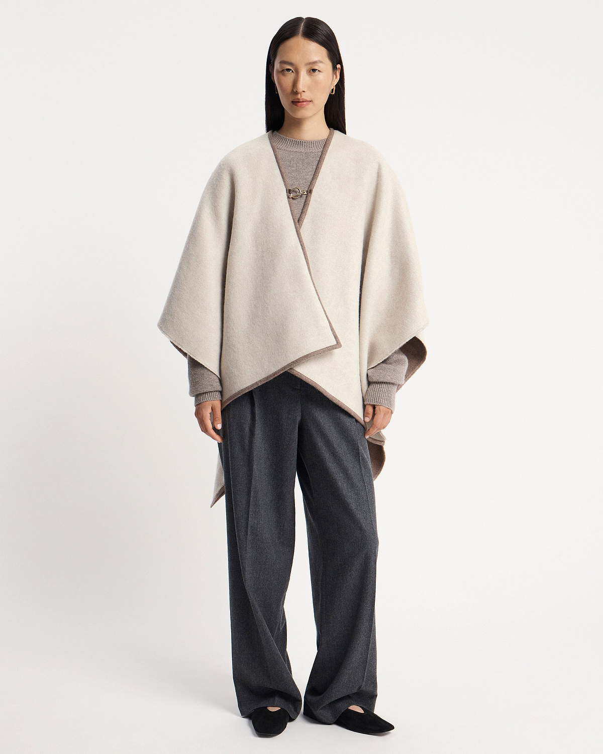 Model wearing a taupe kite woven cape over grey jumper and dark trousers.