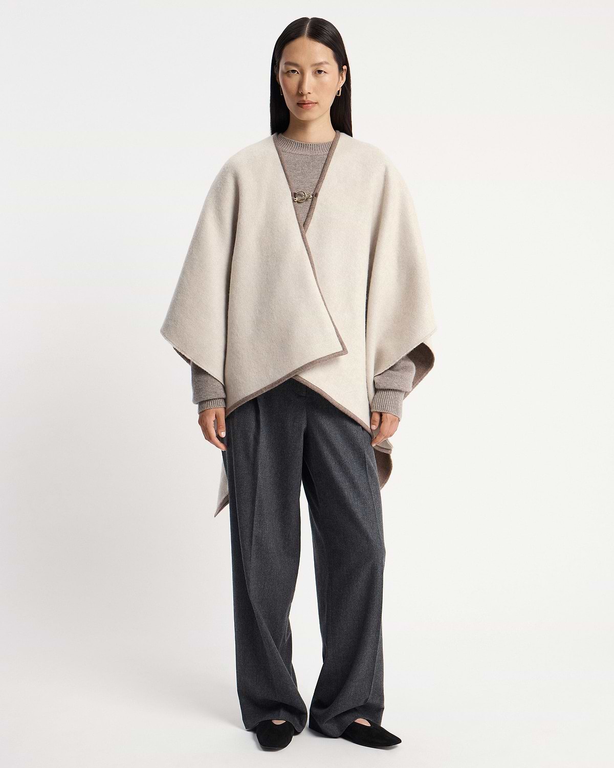 Model wearing a taupe kite woven cape over grey jumper and dark trousers.