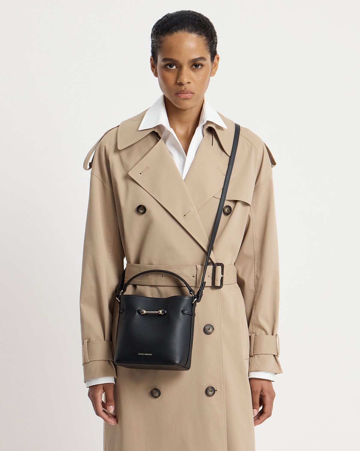 Model wearing a beige trench and black Strathberry Corda Bucket Mini cross-body bag.
