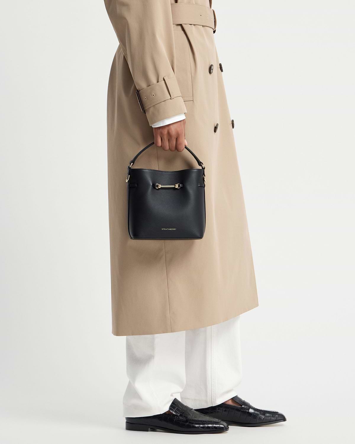 Model holding black Strathberry Corda Bucket Mini bag with beige trench coat.