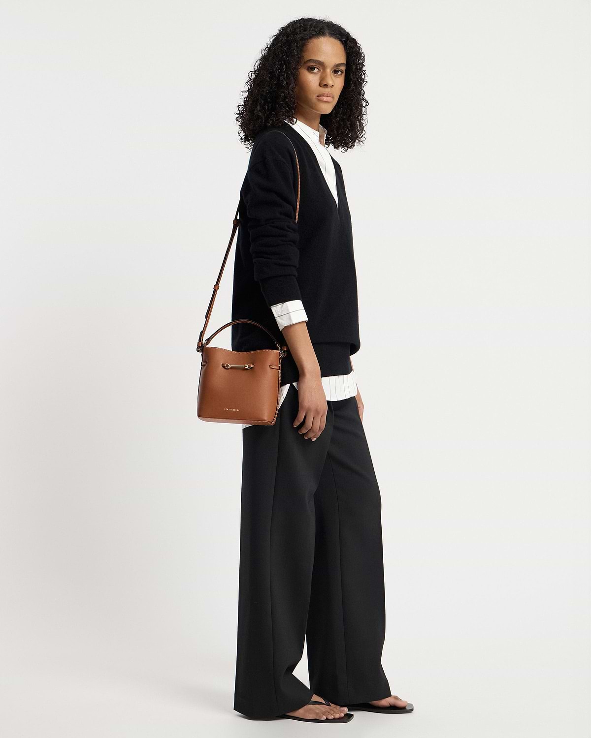 Model wearing Strathberry Corda Bucket Mini chestnut leather bag on shoulder.