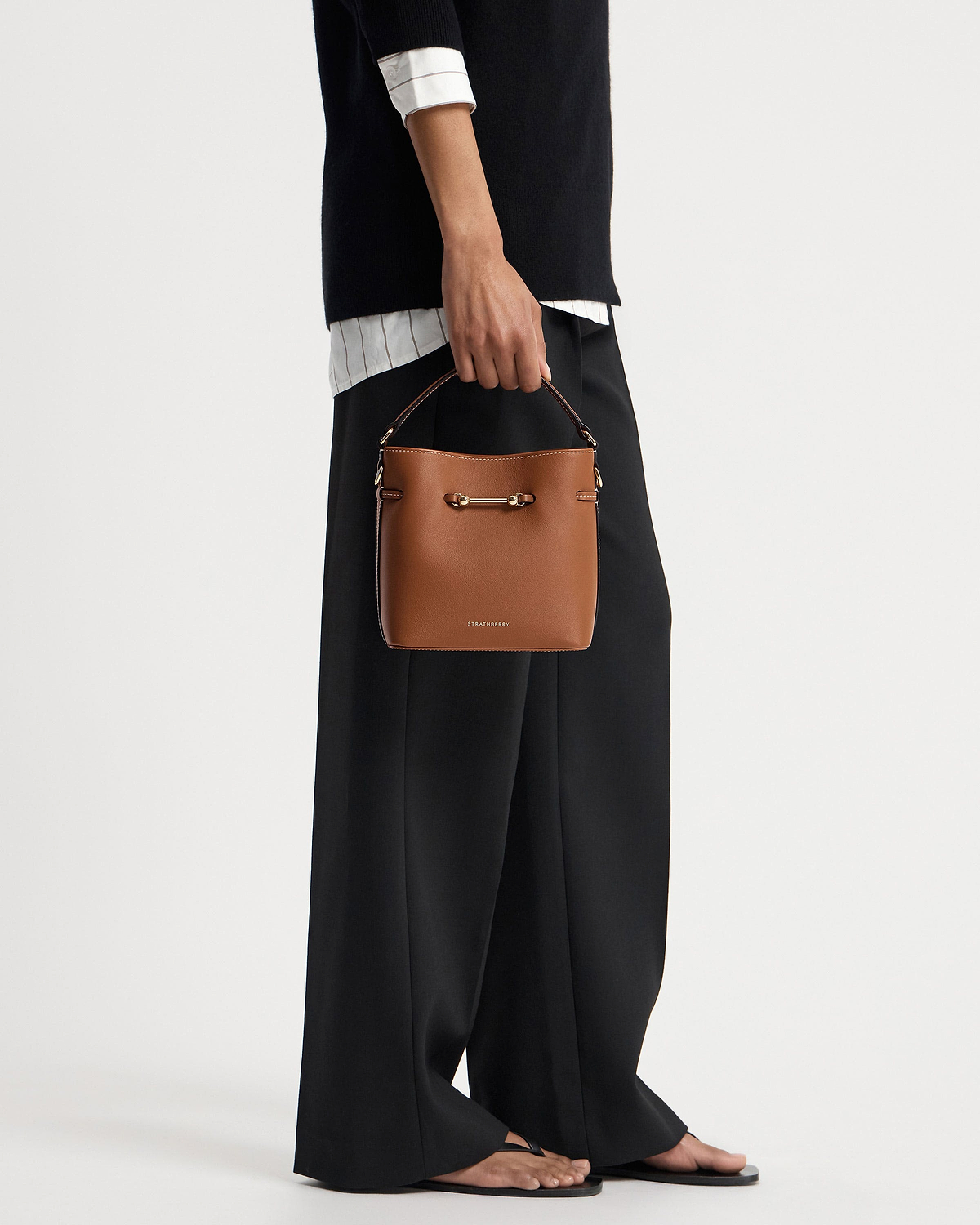 Hand holding chestnut Strathberry bucket bag with gold hardware, model in black trousers.