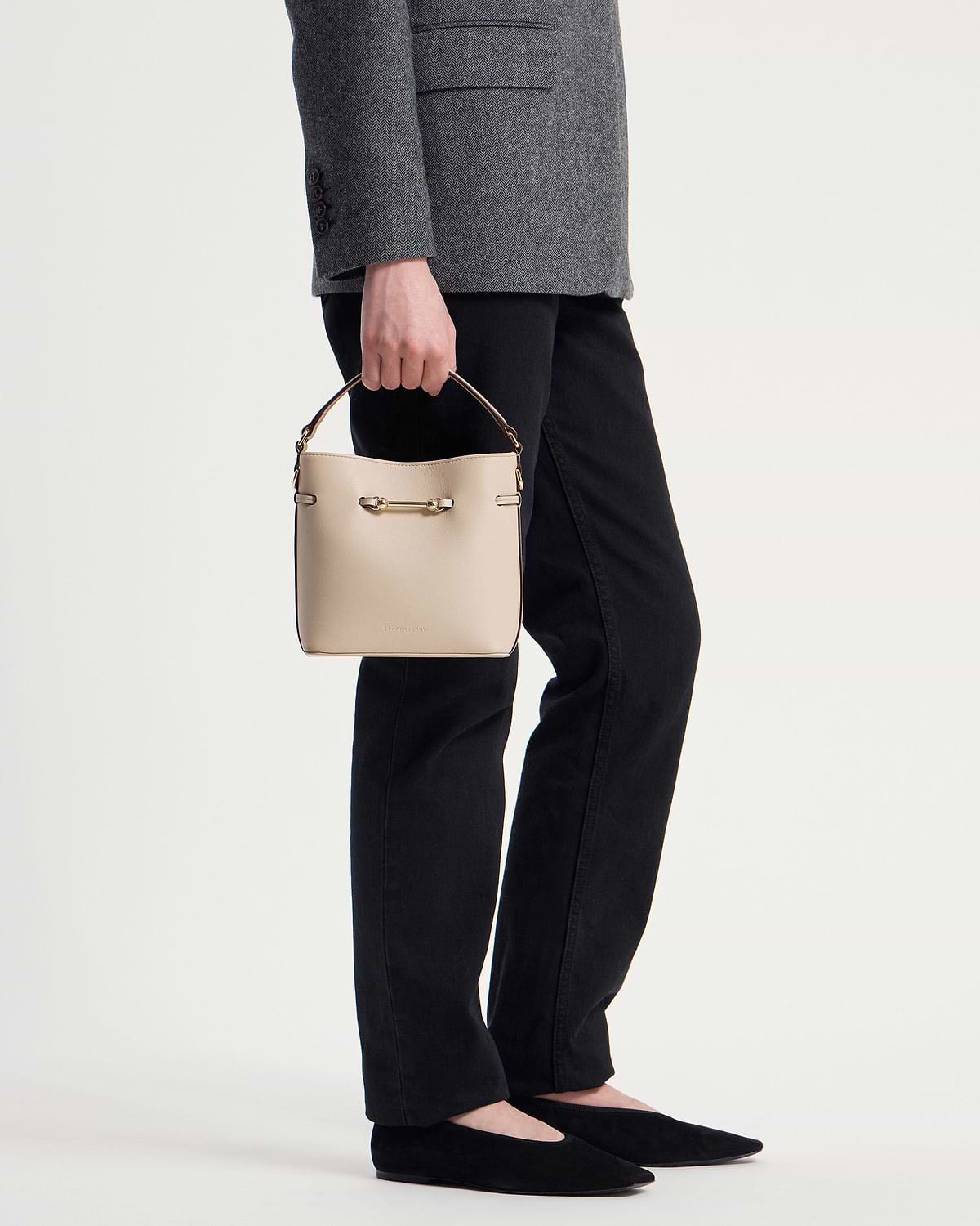 Hand holding an oat‑coloured leather mini bucket bag with top handle