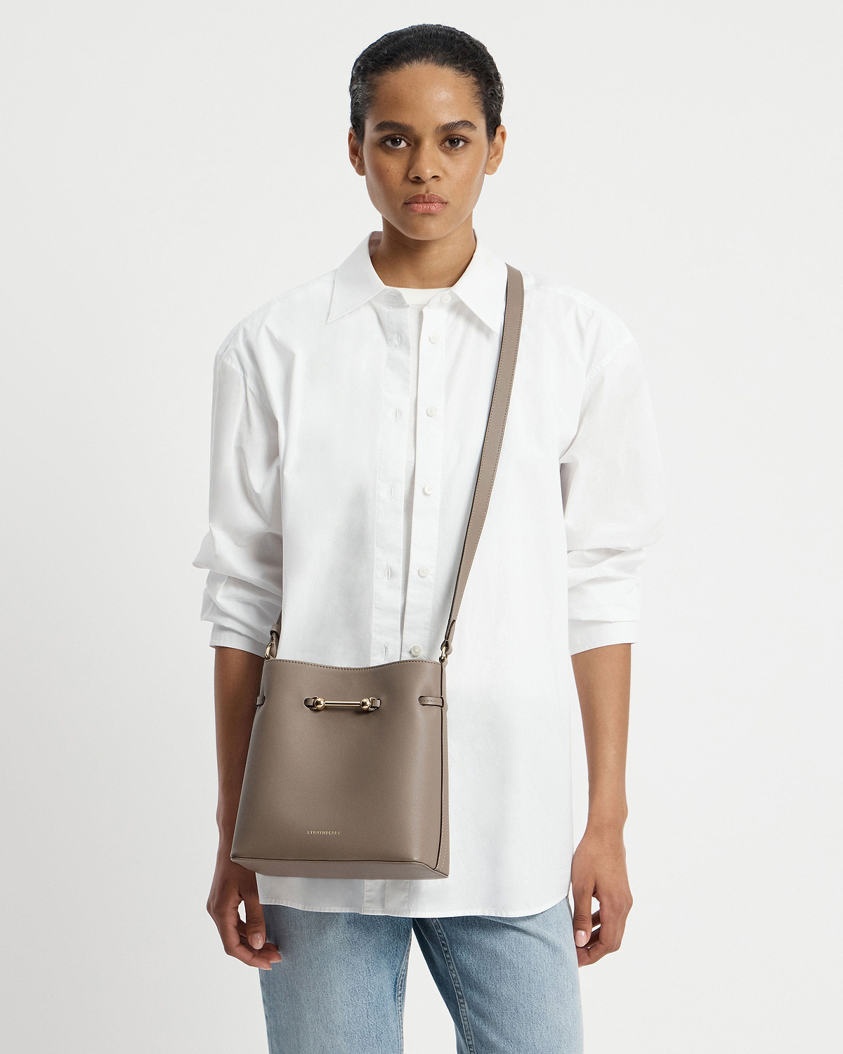 Model wearing white shirt with taupe Corda bucket bag worn crossbody, Strathberry logo visible