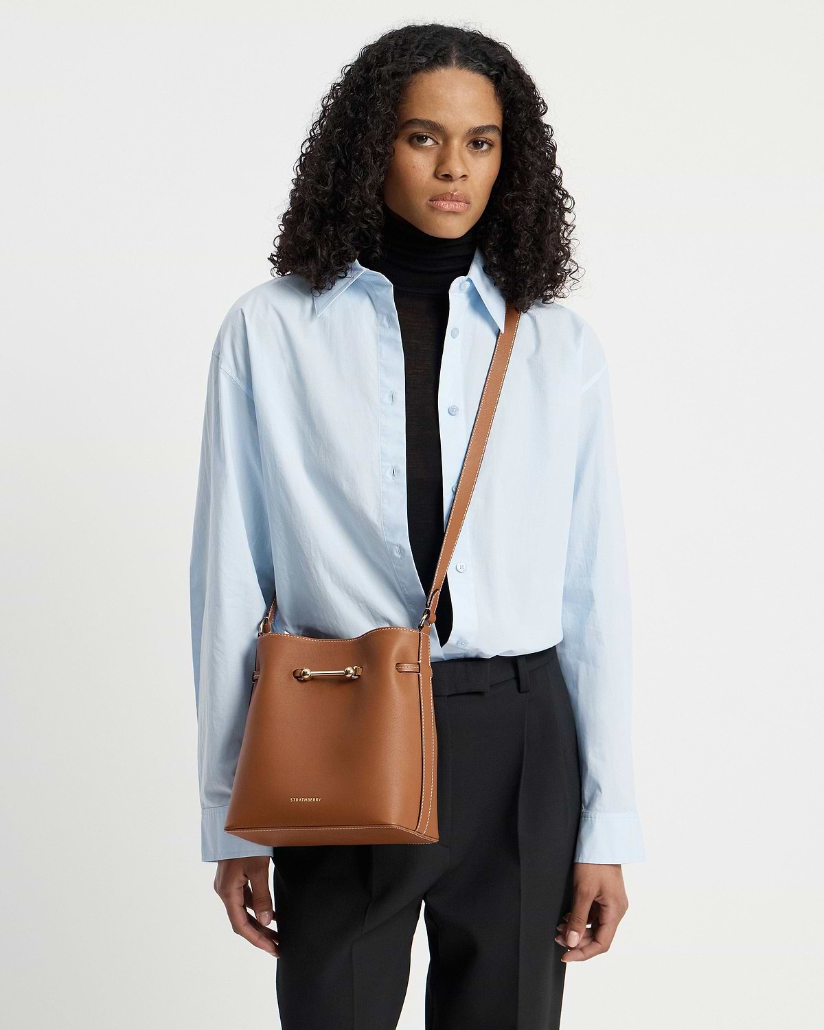 Model wearing chestnut Corda bucket bag by Strathberry, cross-body over blue shirt.