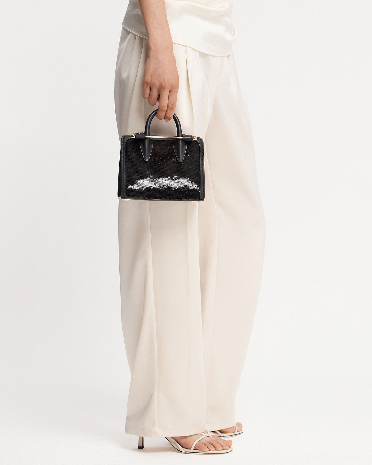 Hand holding black sequinned nano tote with top handles against white trousers.